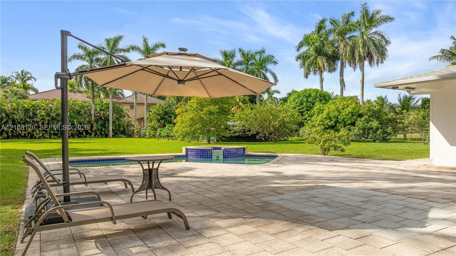 Expansive backyard featuring a stone-paver patio, swimming pool with a blue-tiled raised spillover spa, manicured lawn, and mature palm trees