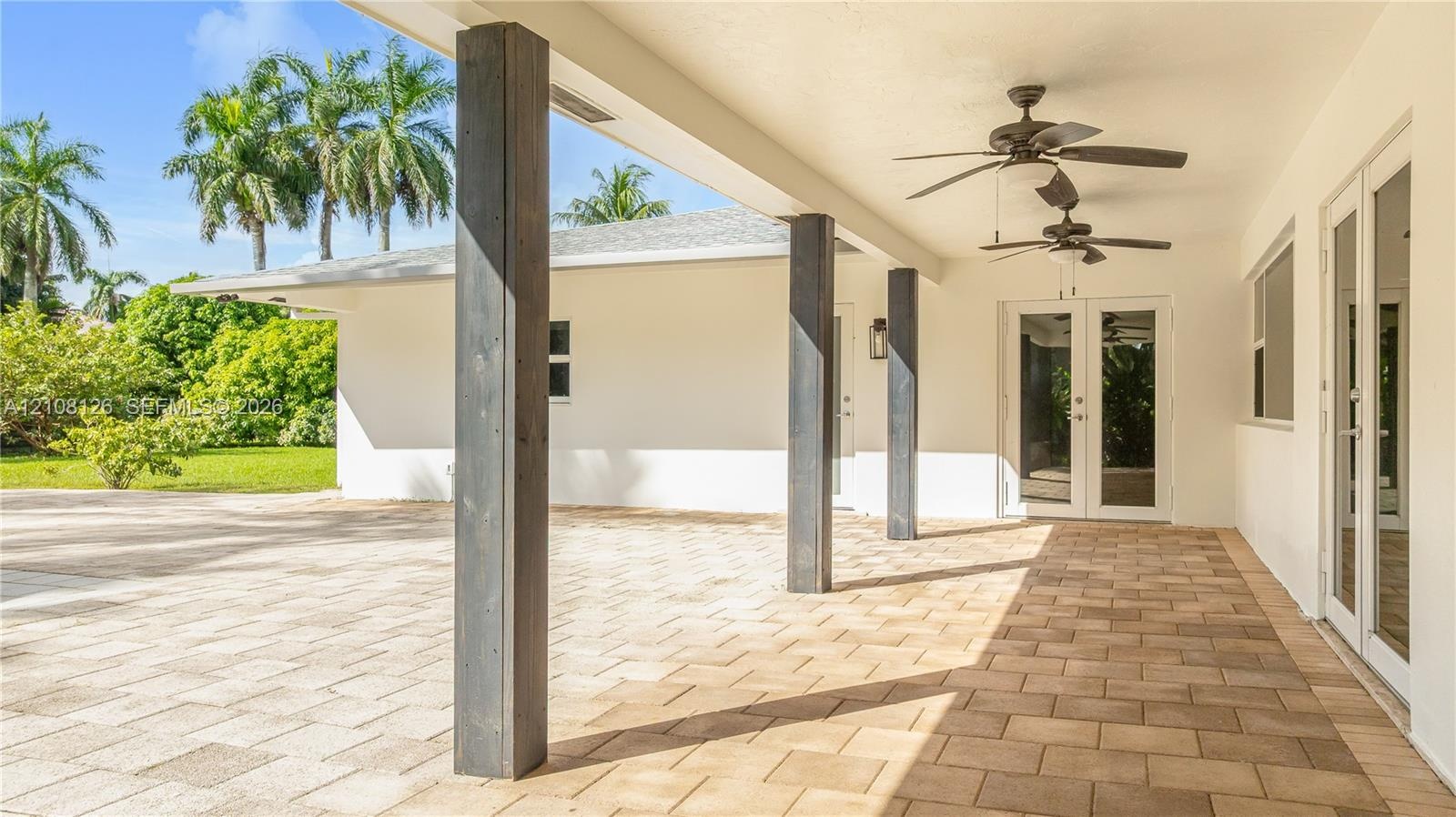 Covered outdoor patio featuring light-toned paver flooring, dark wood-finish structural support columns, dual ceiling fans, and double glass-paned doo