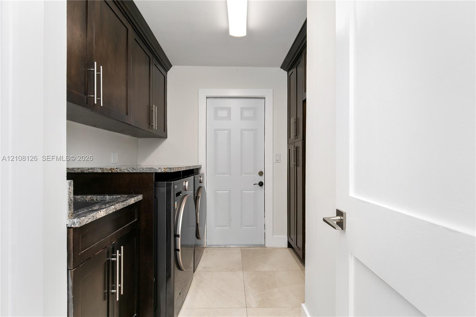 Dark-finish cabinetry with silver-tone hardware, stone-composite countertops, built-in tall pantry storage, and light-tone tile flooring