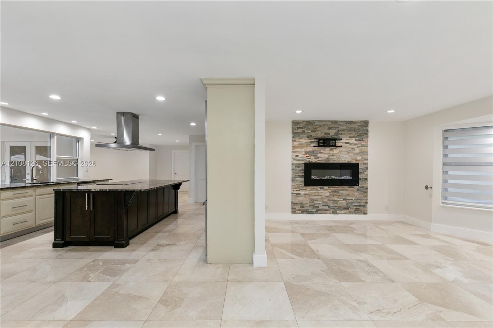 Kitchen featuring a dark-finish island with a countertop, stainless steel range hood, stone-finish fireplace, and neutral-tone tile flooring