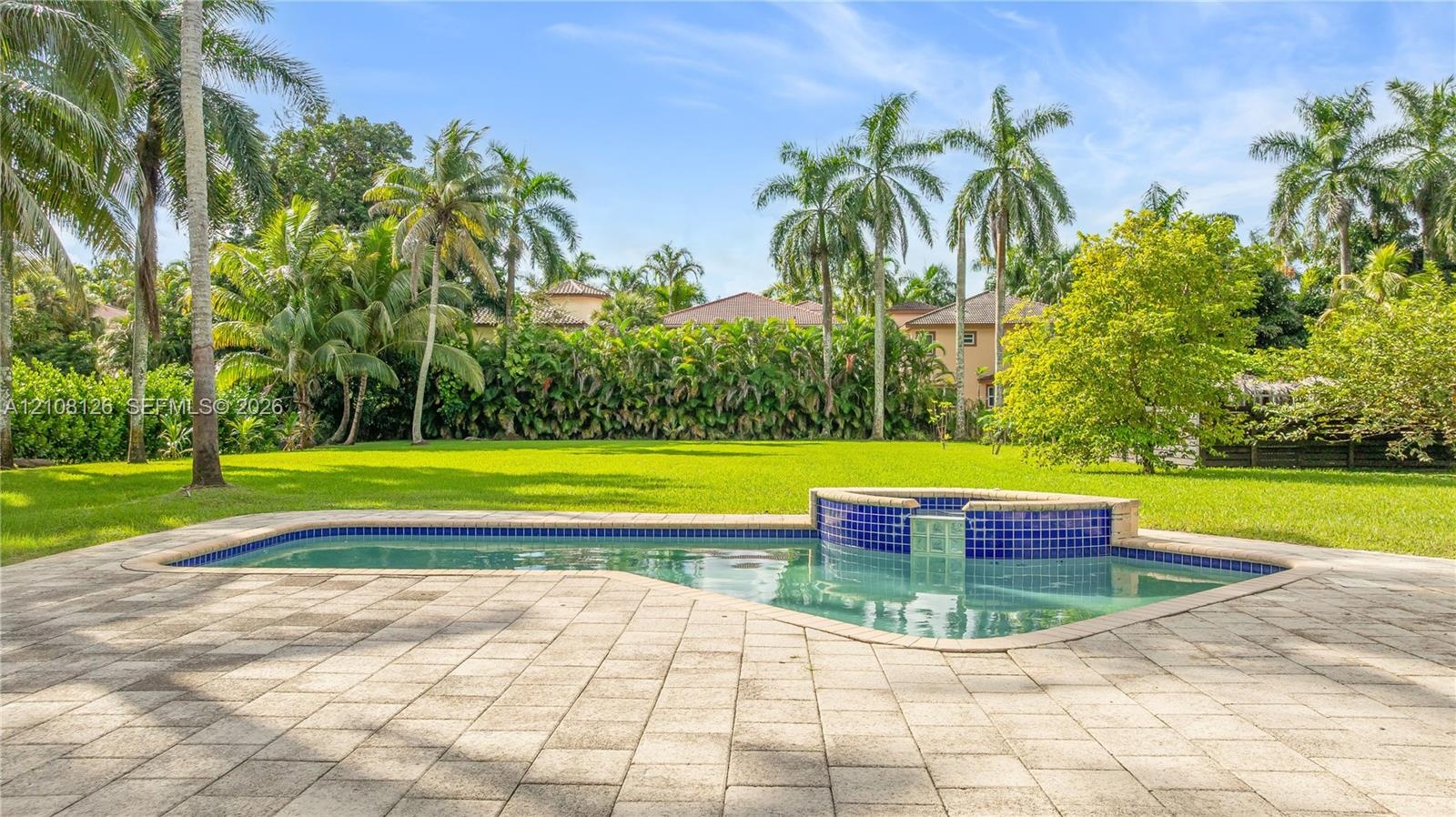 In-ground swimming pool with an attached spillover spa, blue tile detailing, expansive paver patio, and mature palm trees set against a private lawn