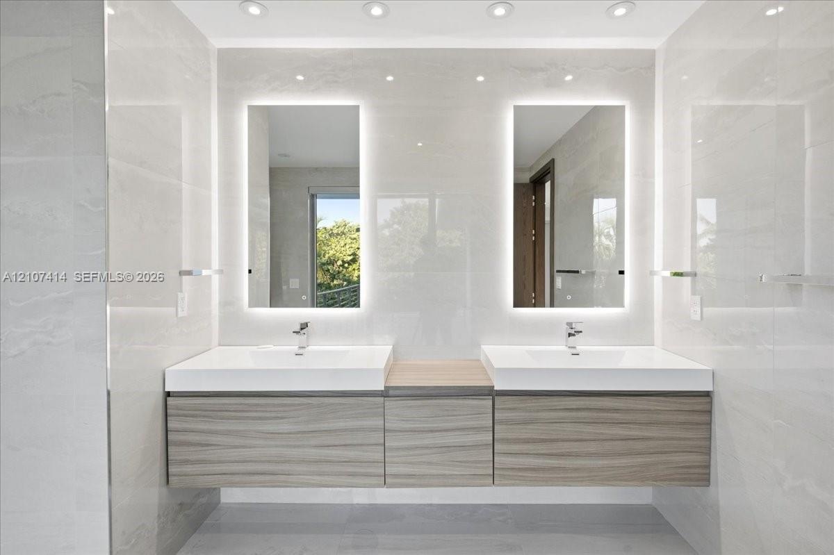 Floating dual vanity with wood-finish cabinetry, white integrated sinks, backlit rectangular mirrors, and recessed ceiling lighting, set against large