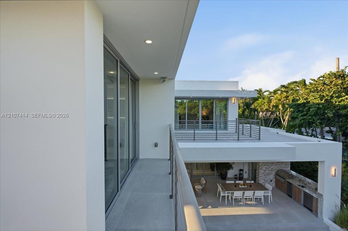 Modern architectural exterior featuring expansive covered balconies, metal railings, recessed lighting, and a ground-level outdoor kitchen with stone 