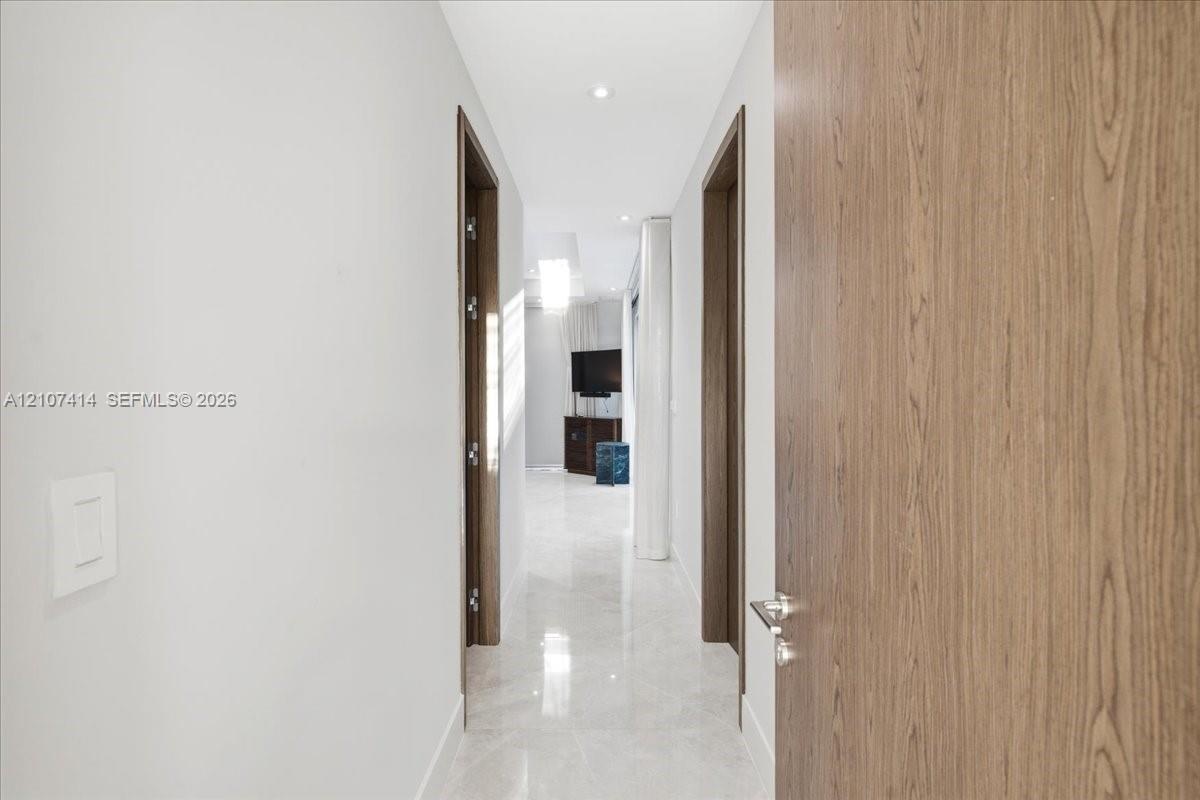Hallway featuring large-format light-toned stone flooring, recessed ceiling lighting, and solid wood-finish doors with brushed-metal hardware