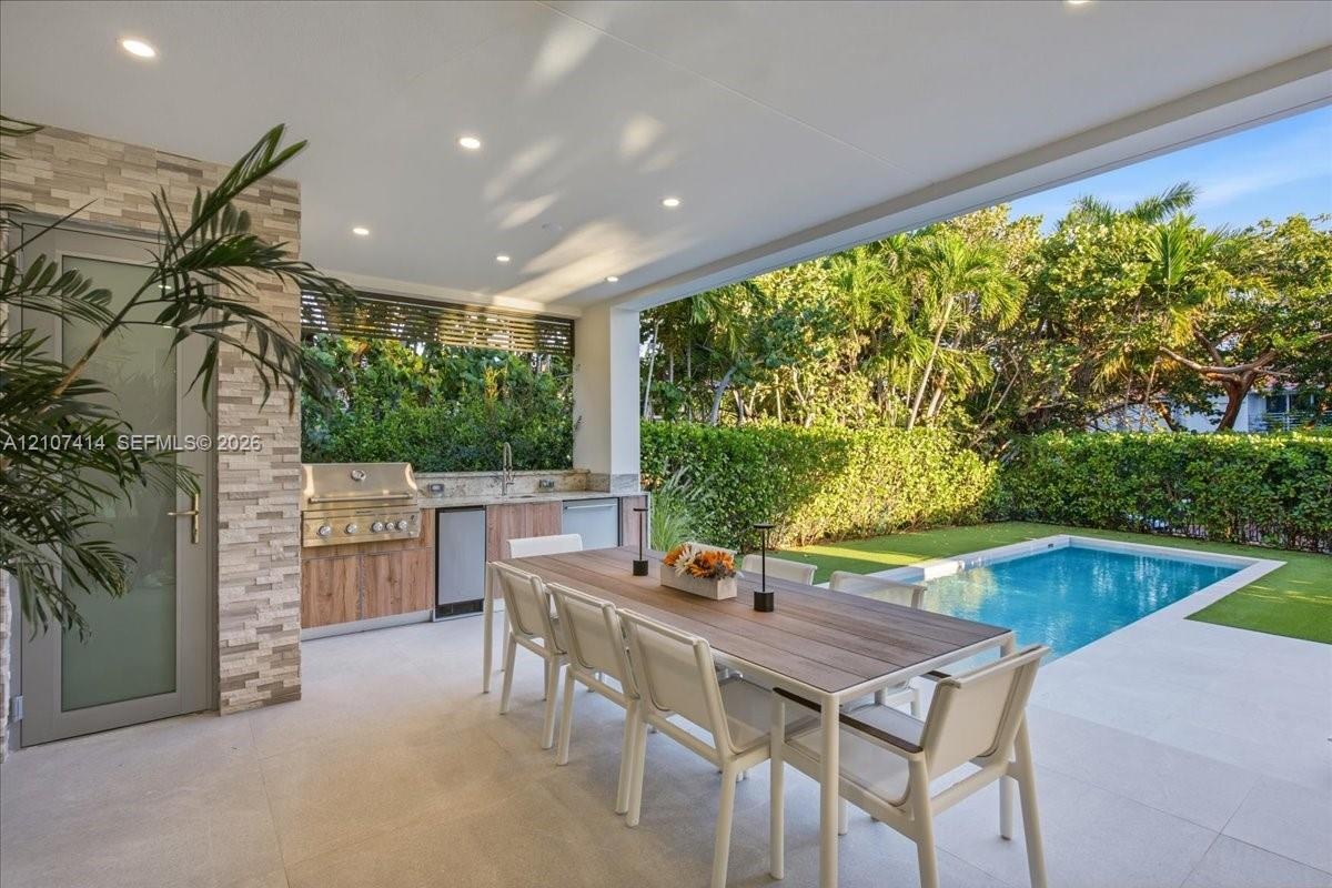 Covered outdoor kitchen featuring stainless steel grill, stone-tile backsplash, and wood-finish cabinetry, complemented by a rectangular pool and ligh