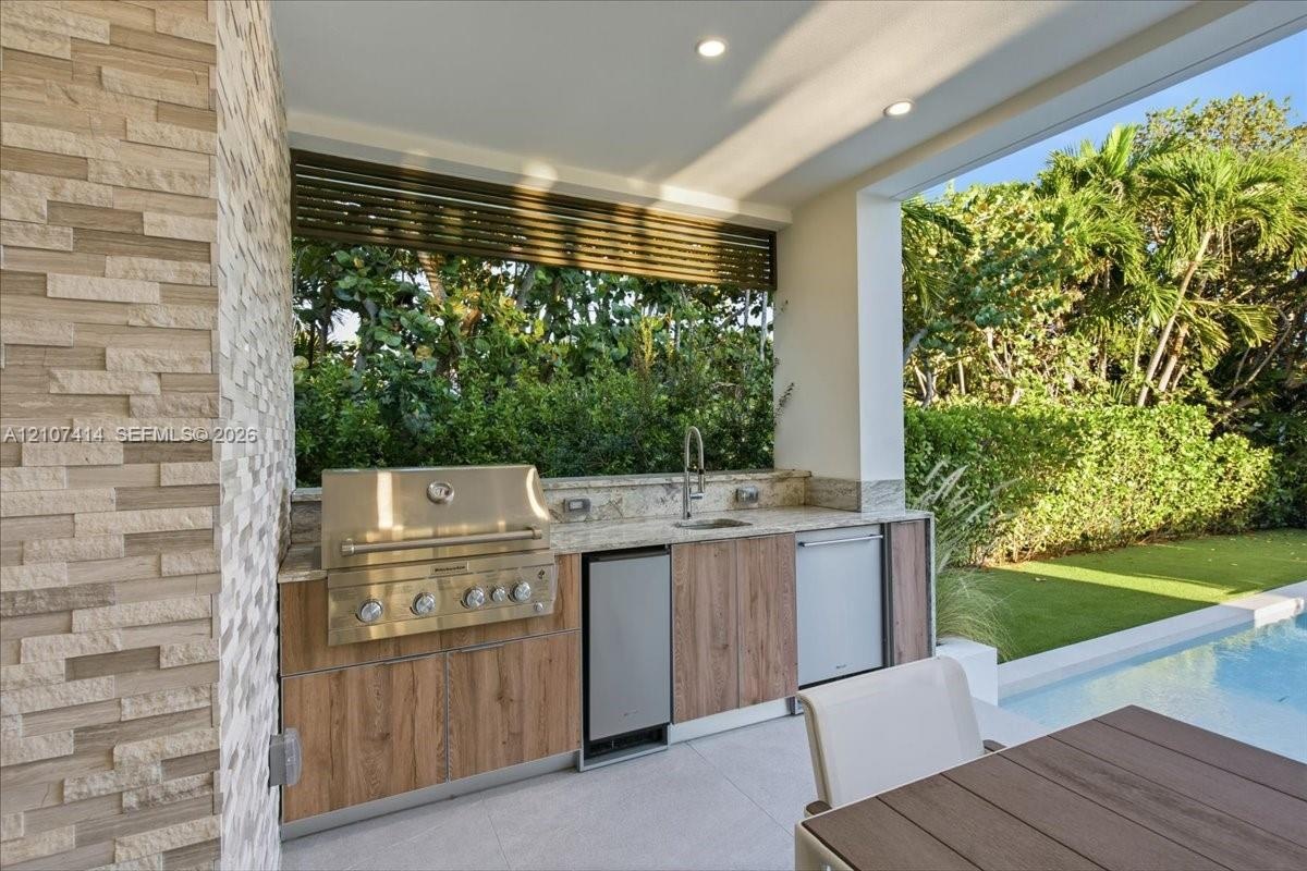 Outdoor kitchen featuring a stone-clad wall, built-in stainless steel grill, stone countertop with integrated sink, wood-finish cabinetry, and recesse