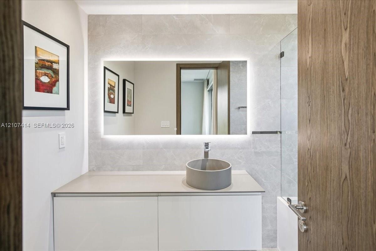 White vanity with a solid-surface countertop, cylindrical vessel sink, LED-backlit mirror, and full-height stone-finish wall tiling