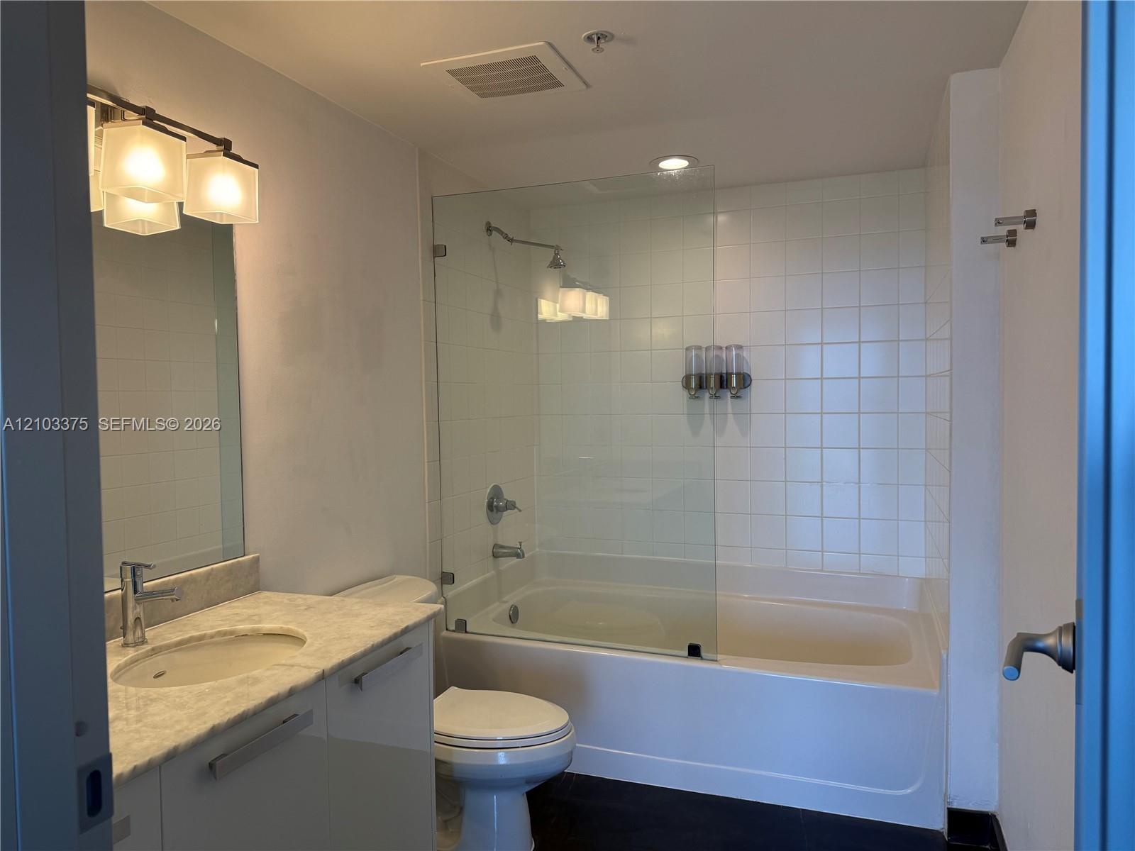 Bathroom featuring a stone-top vanity with an integrated sink, frameless glass shower panel, white tile tub surround, and three-light wall-mounted fix
