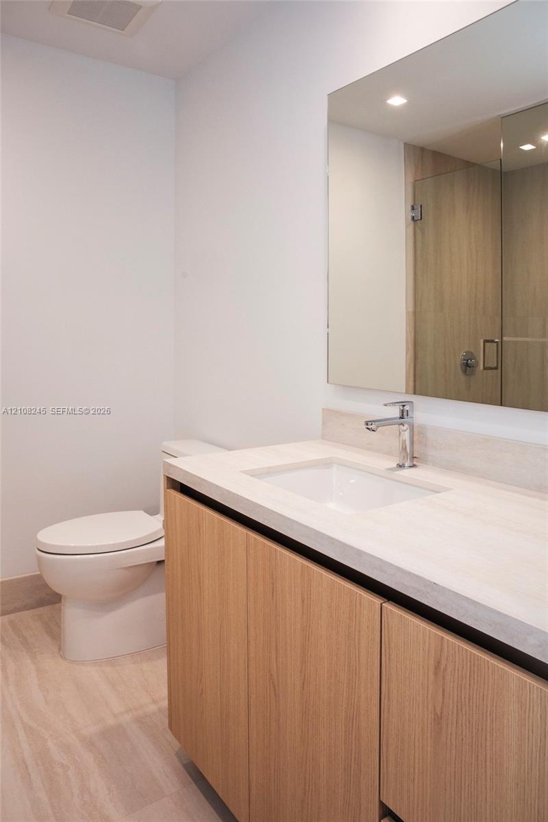 Bathroom featuring a wood-finish vanity with a light-toned countertop, integrated rectangular sink, polished chrome faucet, and a frameless wall mirro
