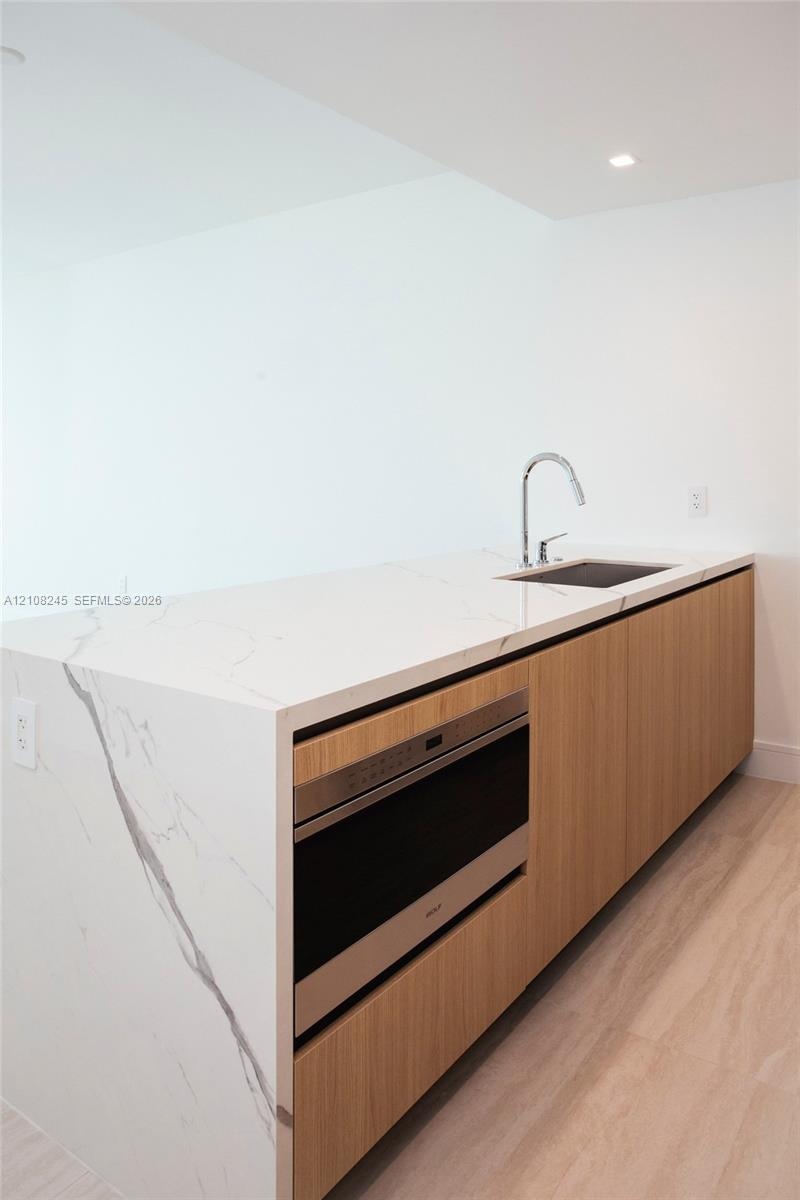 Kitchen island featuring a waterfall countertop with integrated sink and chrome pull-down faucet, flat-panel wood cabinetry, and a built-in microwave 