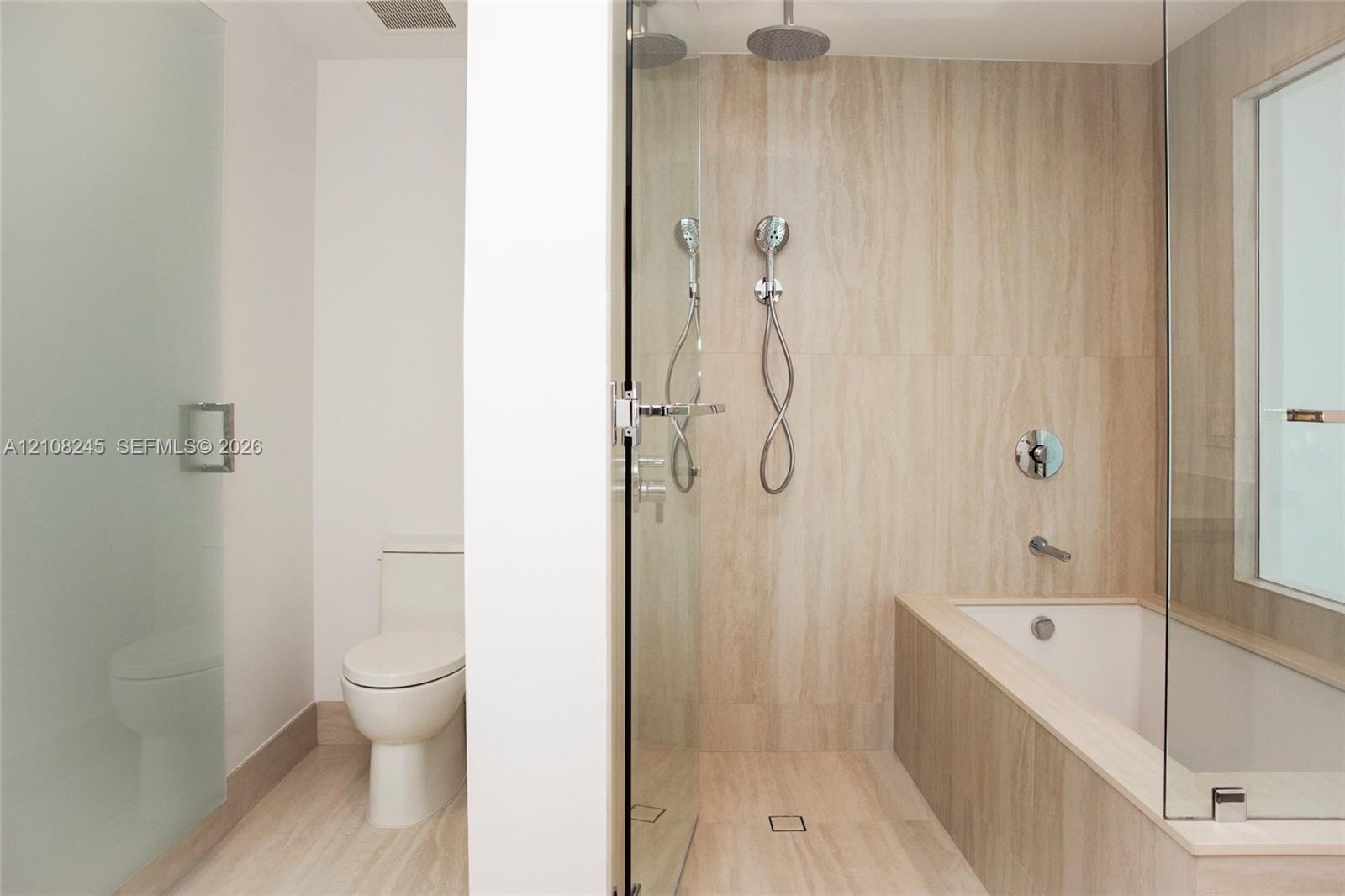 Bathroom featuring stone-finish wall and floor tiling, glass-enclosed shower with rainfall and handheld showerheads, deep soaking tub, and enclosed to