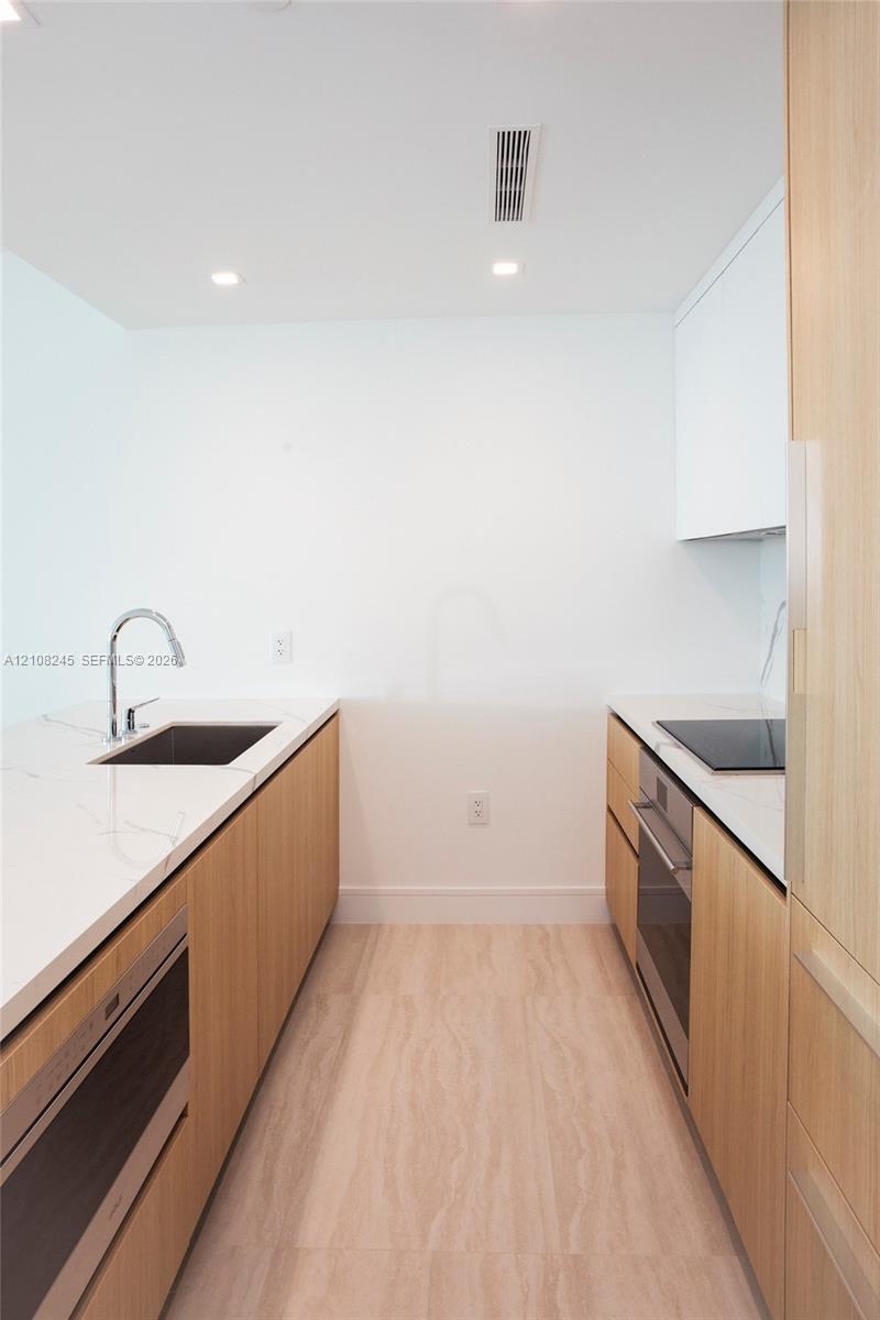Kitchen galley featuring light wood-finish cabinetry, white stone-finish countertops, an undermount sink with a gooseneck faucet, a built-in oven, and