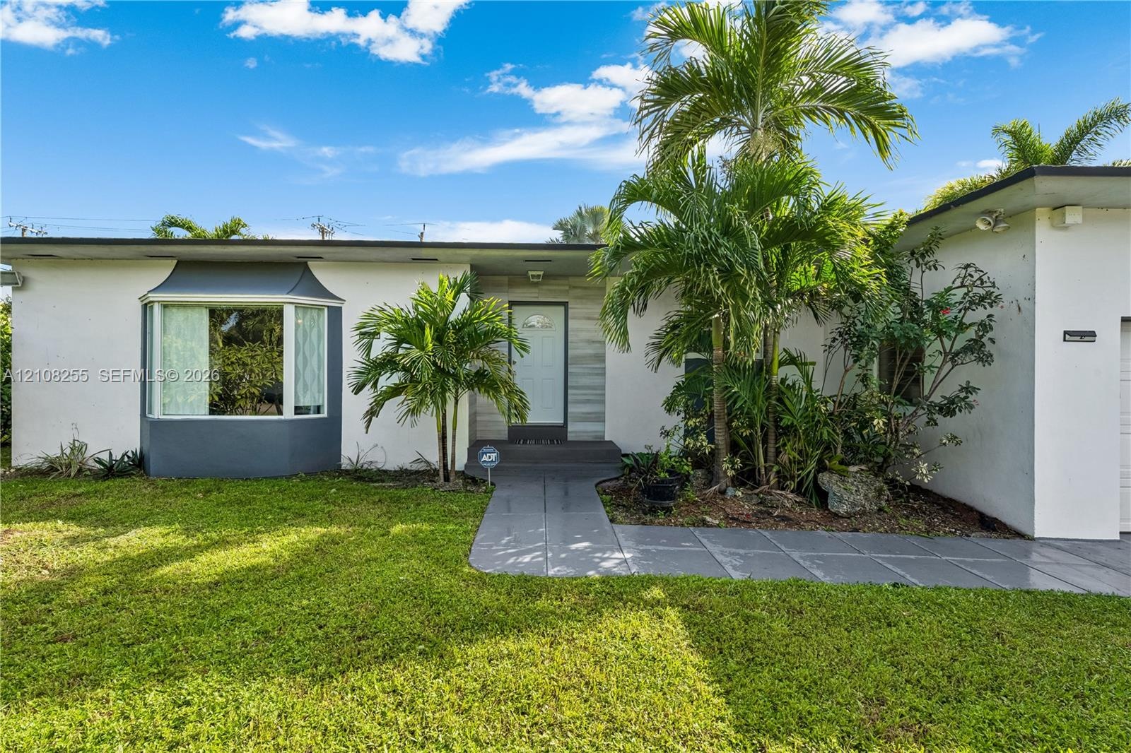 Single-story exterior featuring white stucco finish, front bay window, stone-accented entryway, gray concrete walkway, and established palm landscapin