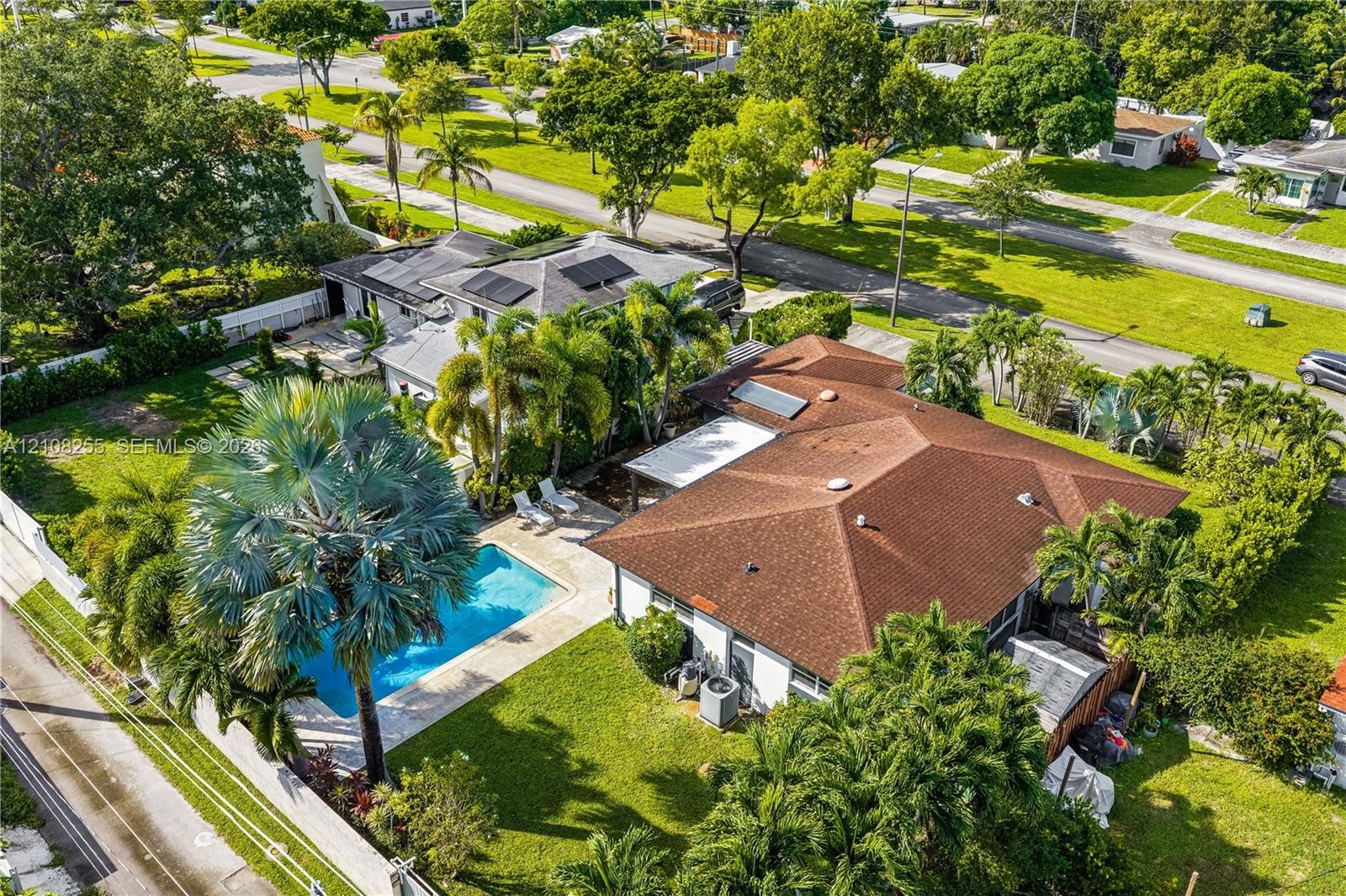 Aerial perspective of a property featuring an in-ground swimming pool, a patio area with a pergola, roof-mounted solar panels, and mature palm landsca