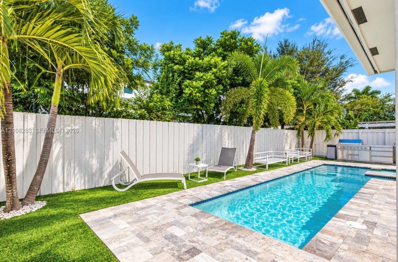 Private backyard featuring a rectangular swimming pool, stone paver decking, synthetic turf, mature palm trees, and a built-in outdoor kitchen