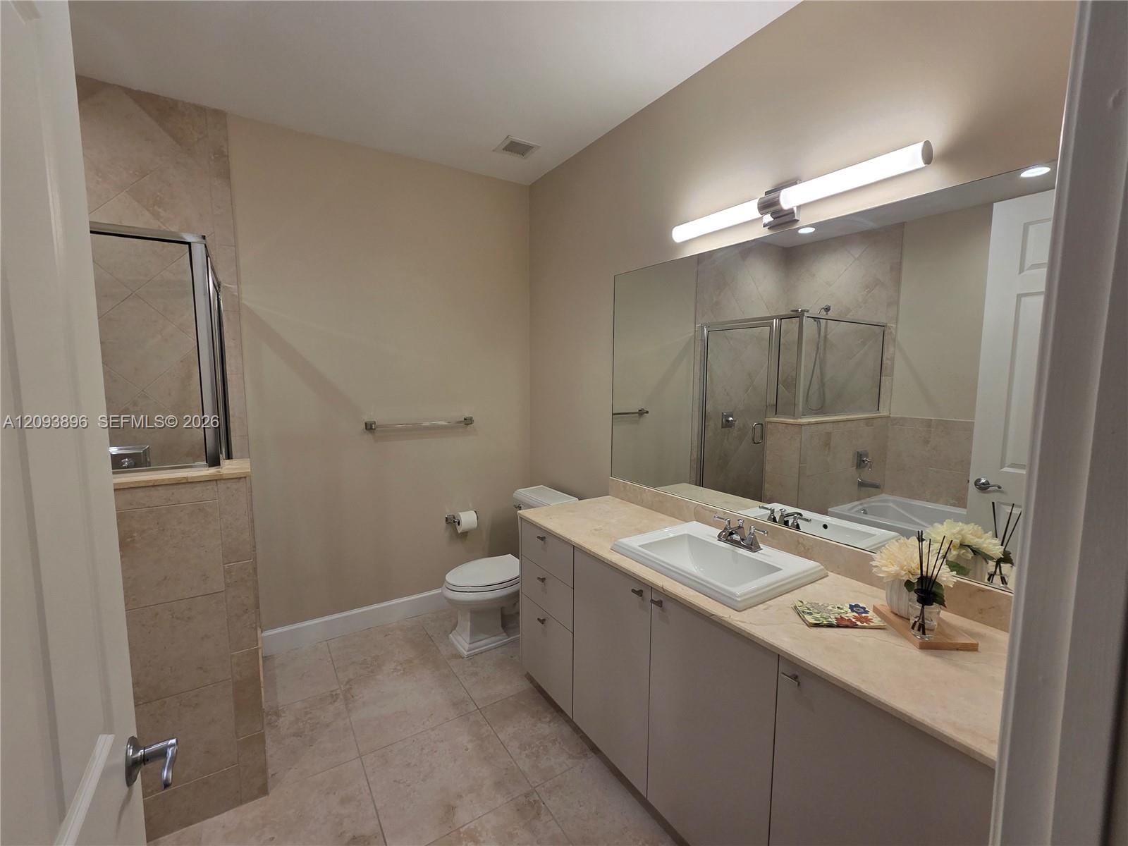 Bathroom featuring a vanity with integrated sink, stone-surface countertop, full-width wall mirror, tile flooring, and walk-in shower with tile surrou