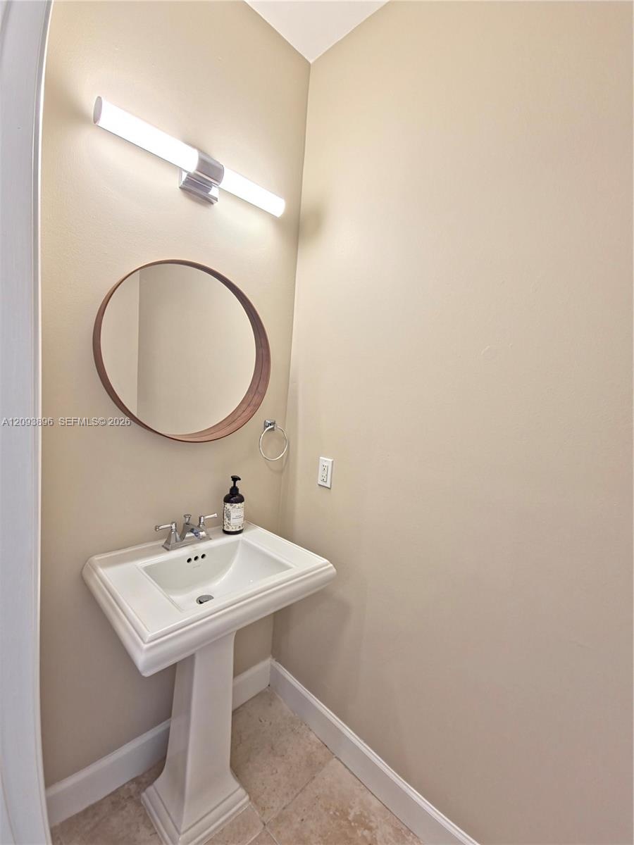 Pedestal sink featuring a chrome faucet, round wall-mounted mirror with a wood-tone frame, linear vanity light fixture, and stone-tile flooring