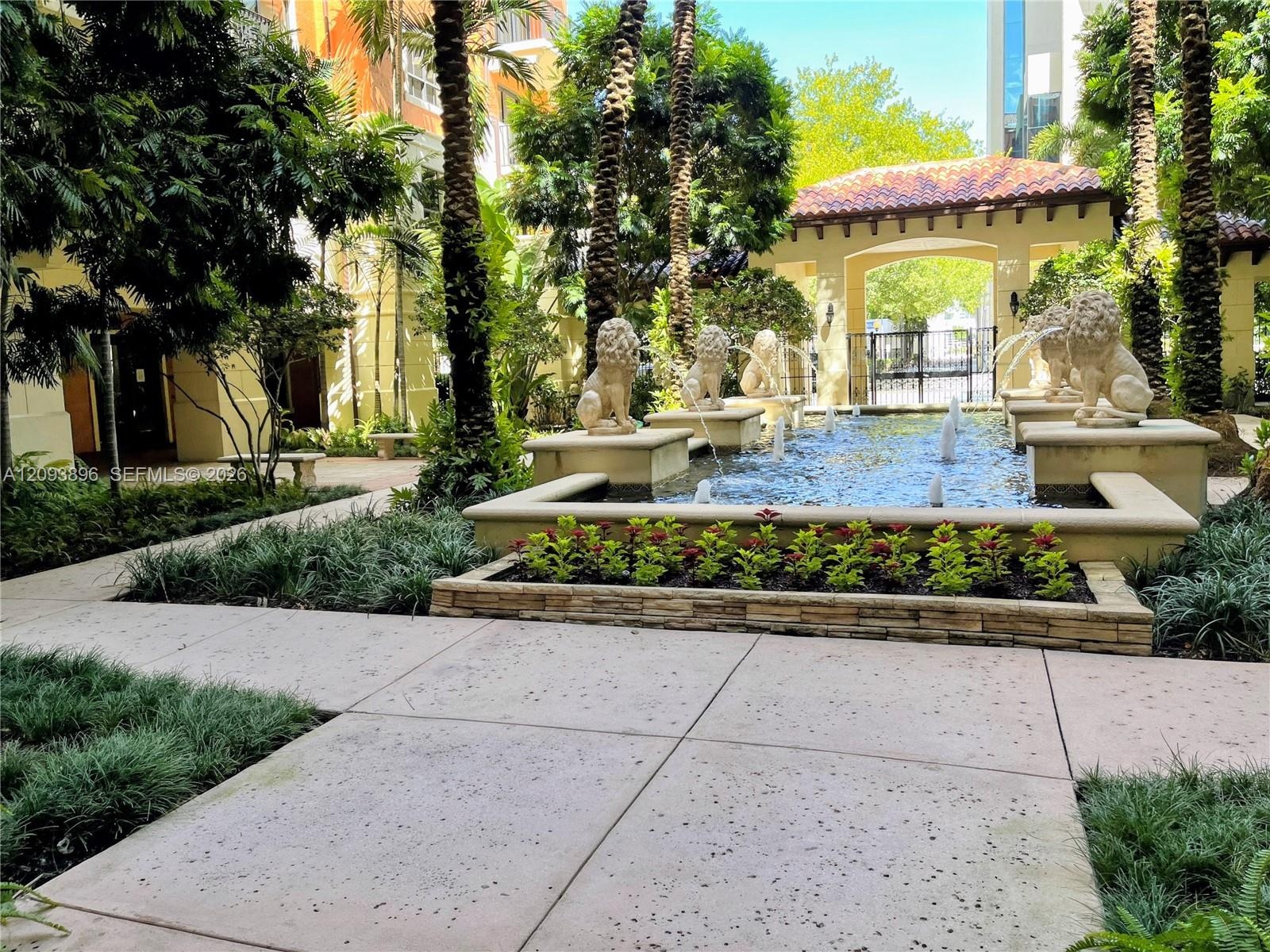 Courtyard featuring a tiered fountain with stone lion sculptures, concrete walkways, mature landscaping, and a covered architectural archway