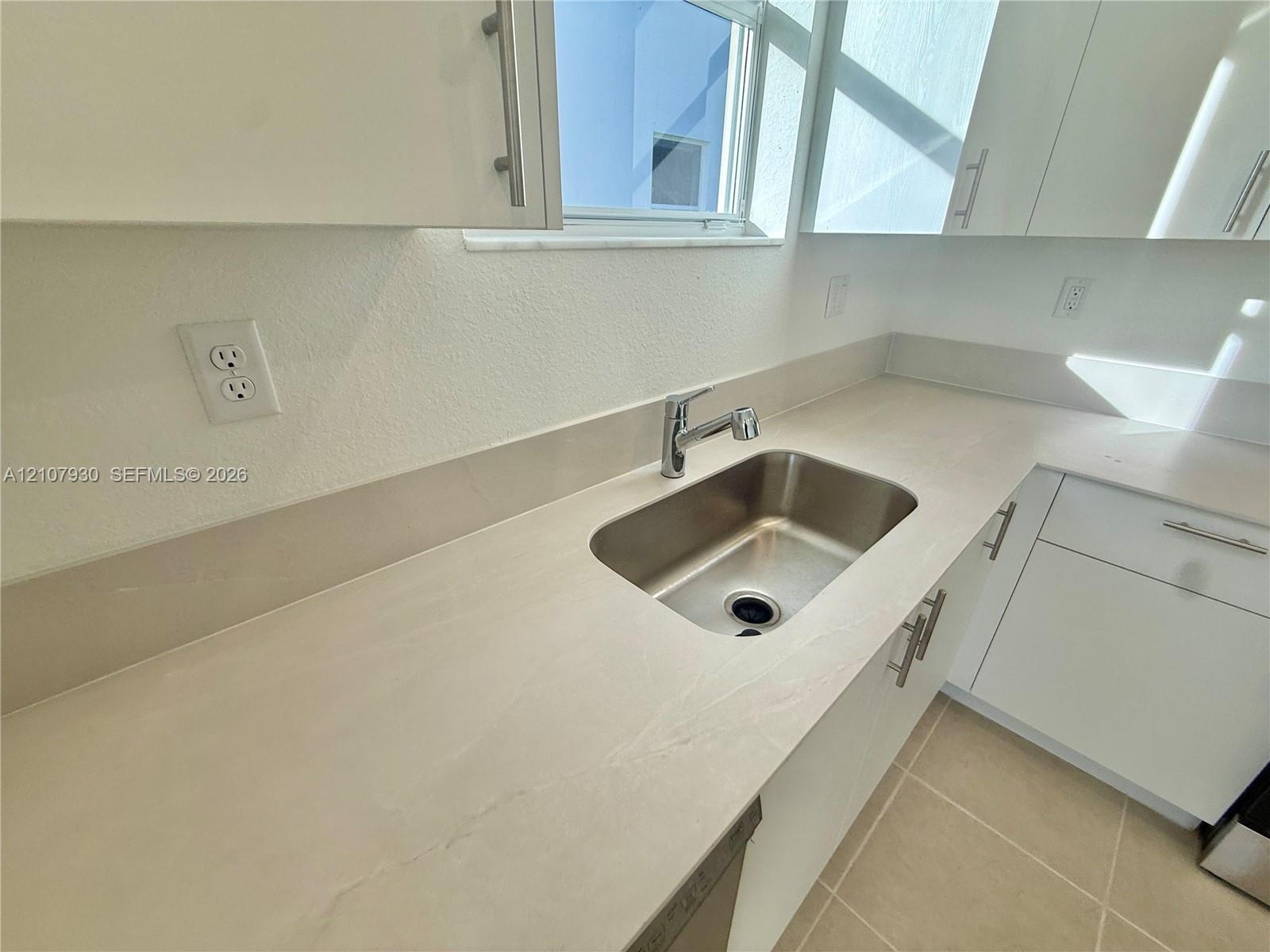 Kitchen featuring white cabinetry with brushed metal hardware, solid-surface countertops, an integrated stainless steel under-mount sink, and ceramic 