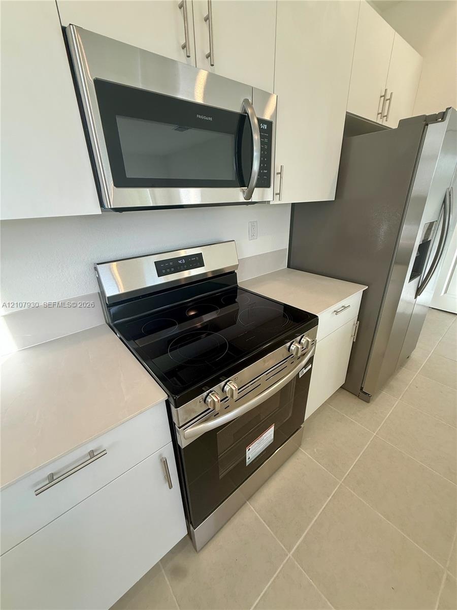 Kitchen featuring flat-panel white cabinetry, stainless steel microwave, glass-top electric range, stainless steel refrigerator, and neutral-toned flo