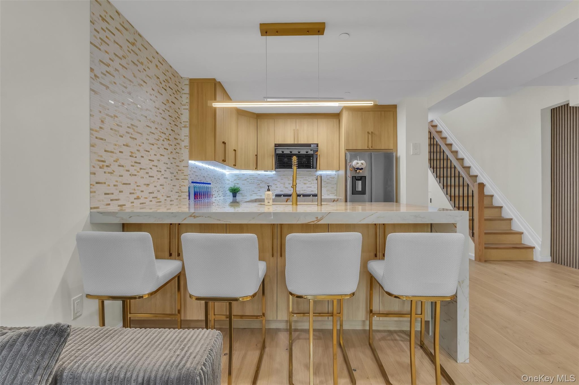 Kitchen featuring light wood-finish cabinetry, waterfall-edge stone countertop, decorative tile backsplash, integrated stainless steel appliances, and