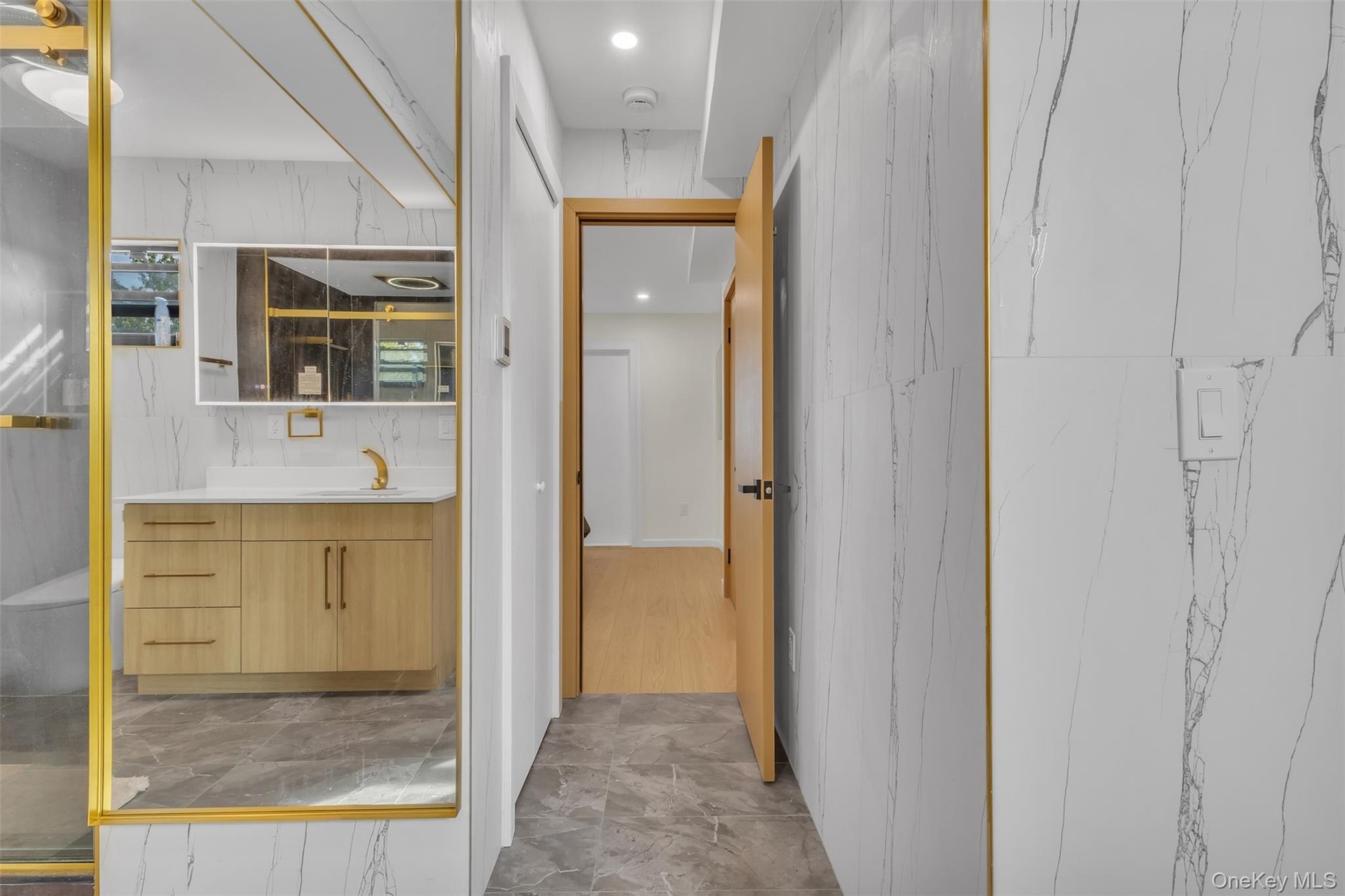 Bathroom featuring large-format wall tiles with veining, wood-finish cabinetry with metallic hardware, recessed lighting, and a gold-trimmed mirror
