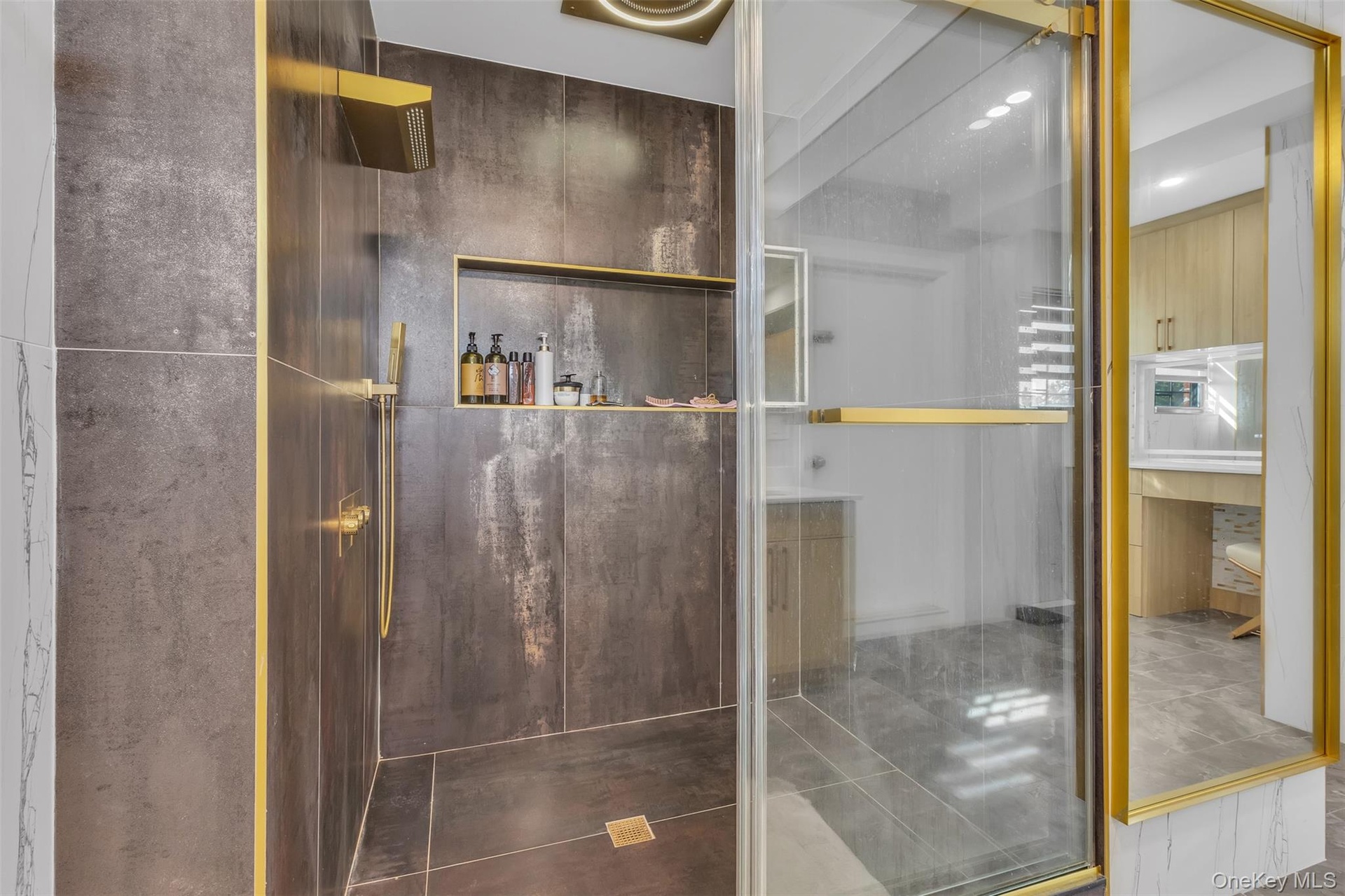 Walk-in shower featuring large-format metallic-finish wall tiles, built-in recessed shelving, gold-tone hardware, and a frameless glass door enclosure