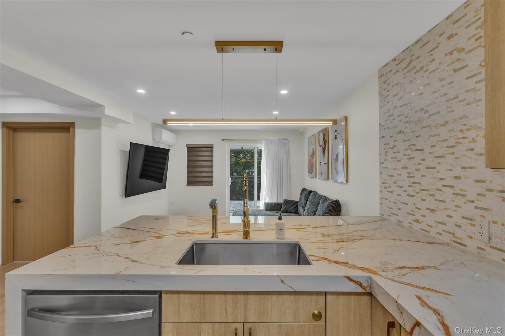 Kitchen island with stone-finish countertop, undermount stainless steel sink, gold-tone faucet, linear pendant lighting, and textured tile backsplash