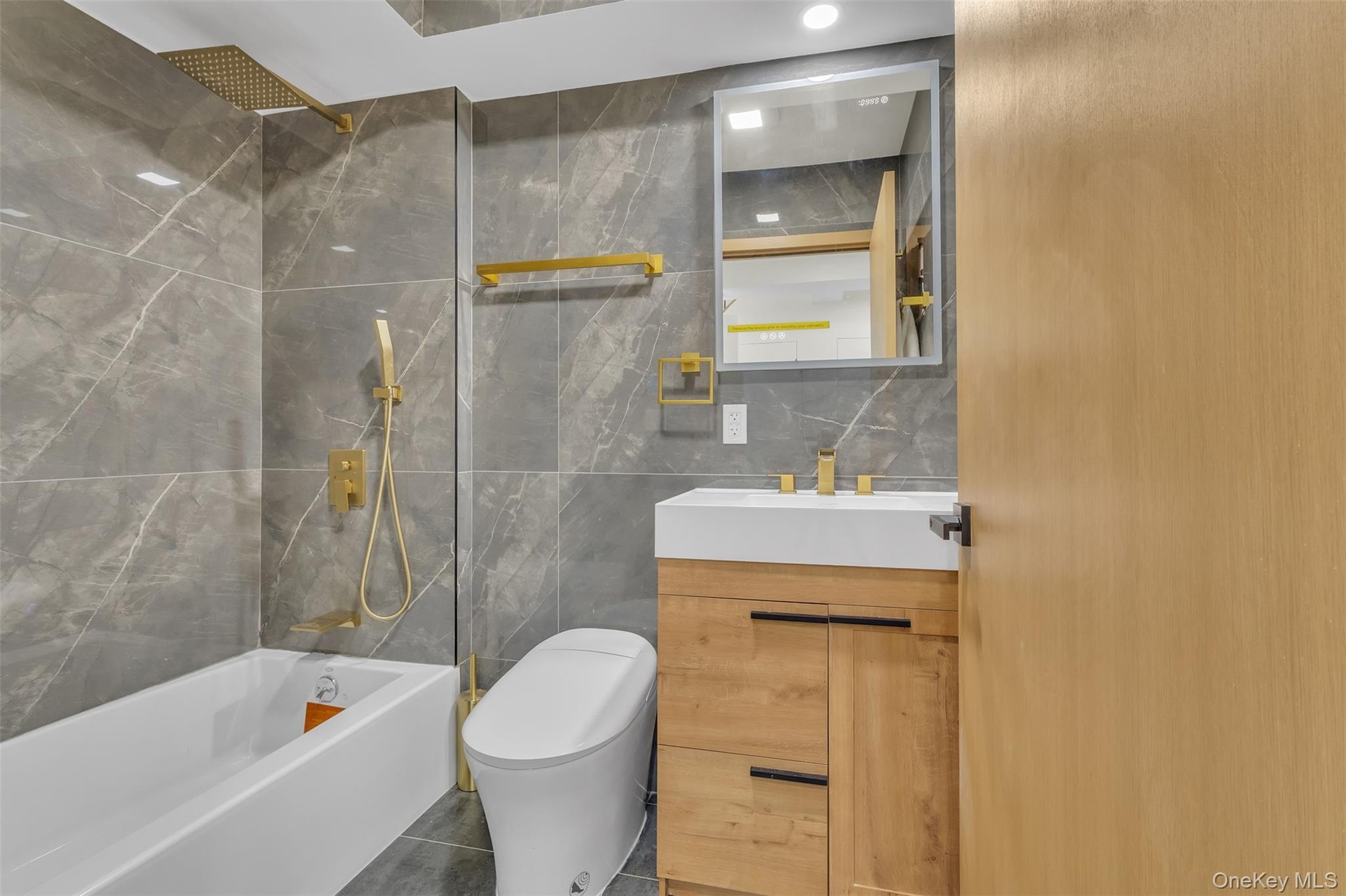 Bathroom featuring wall-to-ceiling large-format tile, vanity with wood-finish cabinetry, brass-tone plumbing fixtures, wall-mounted mirror, and a buil
