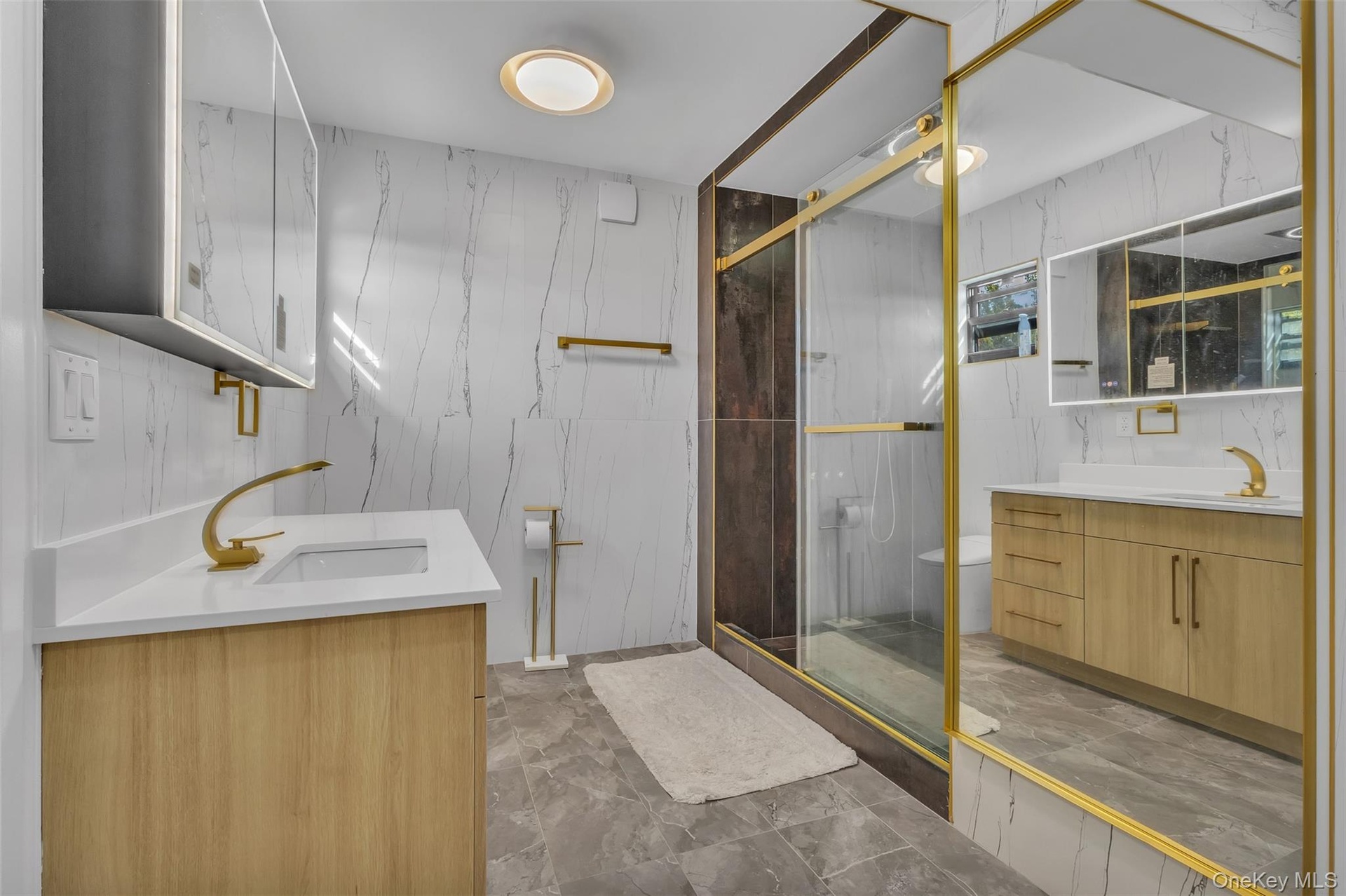 Bathroom featuring wood-finish cabinetry, white solid-surface countertops, gold-toned hardware, full-height tile wall cladding, and a glass-enclosed s