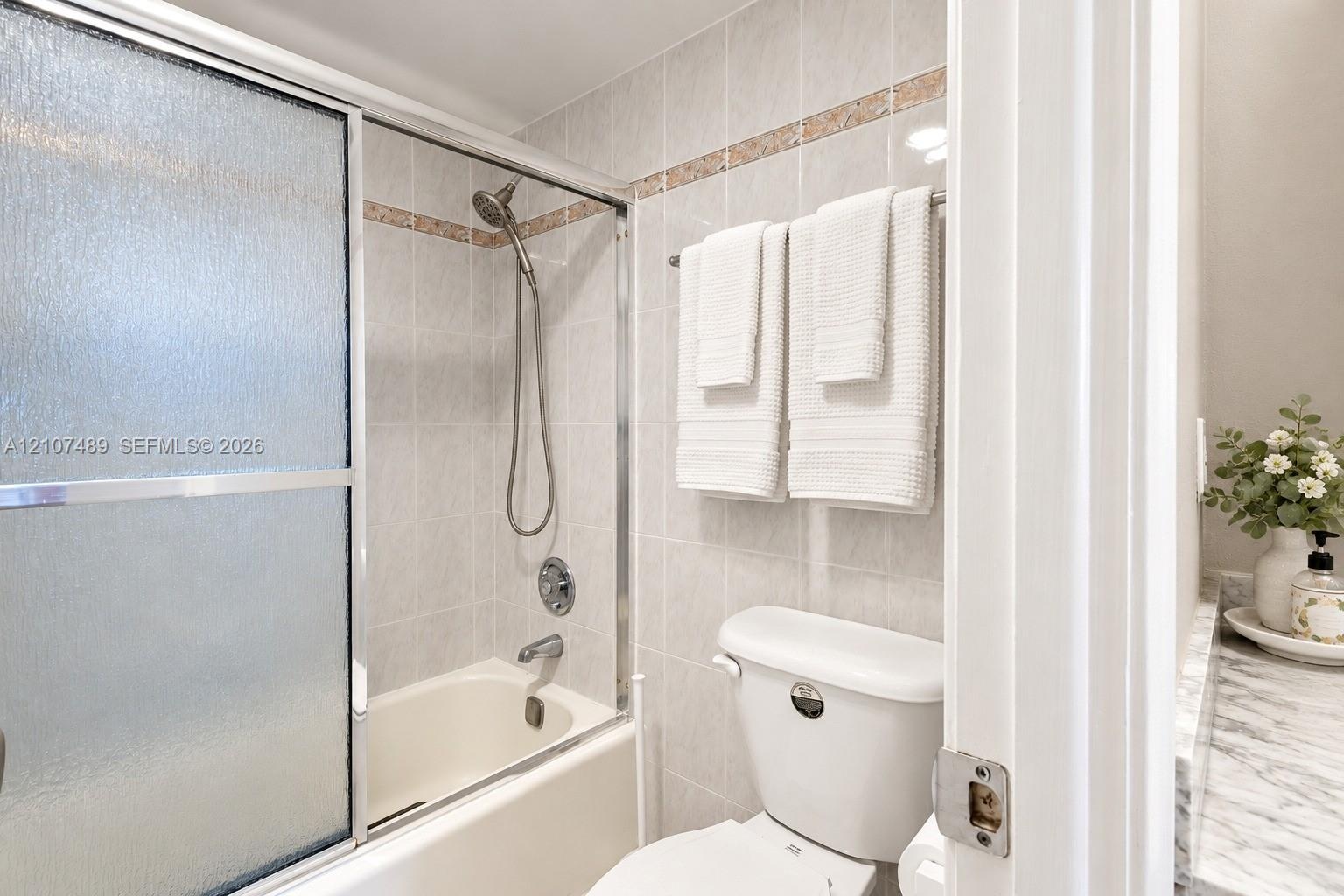 Bathtub with glass sliding doors, integrated handheld shower fixture, light-toned wall tile with decorative accent border, and stone-finish vanity cou