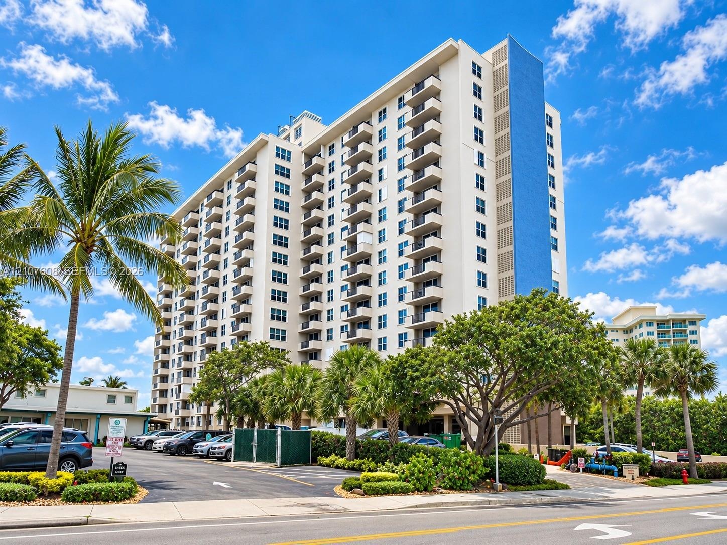 High-rise residential exterior featuring stacked private balconies, a distinctive vertical blue facade accent, and paved surface parking framed by mat
