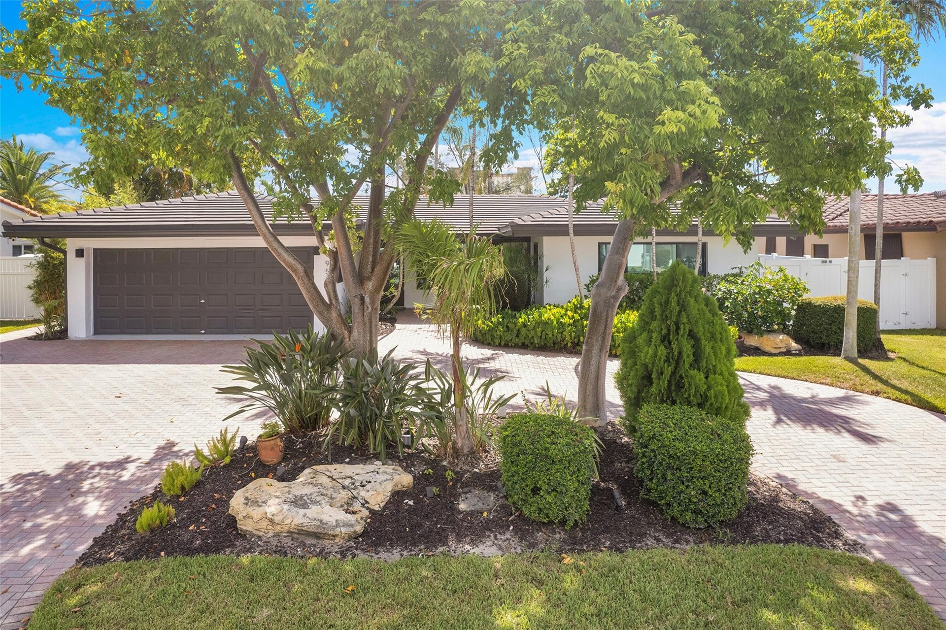 Paver driveway, attached garage, lush landscaping, mature trees, and a white exterior
