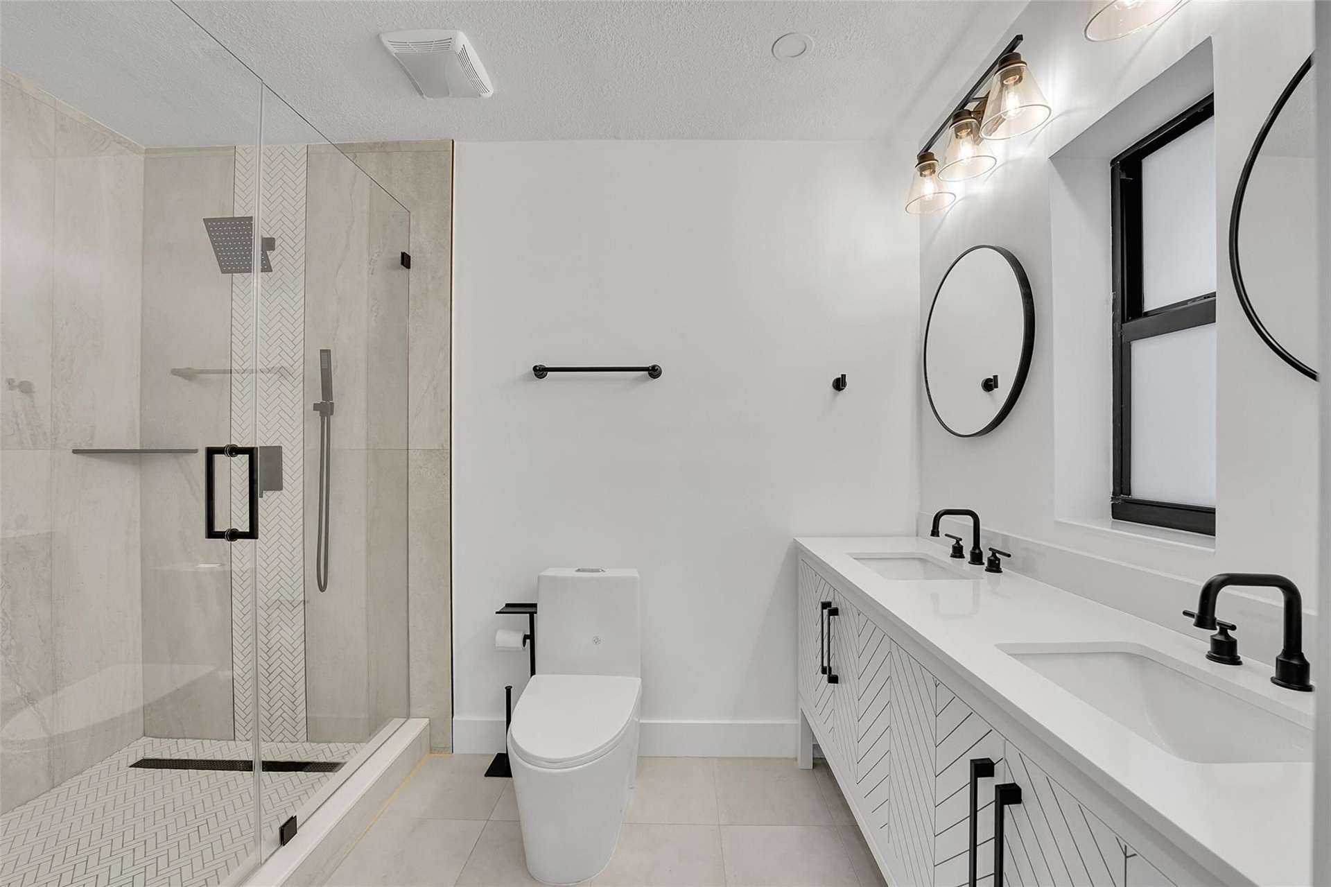 Dual vanity with integrated sinks and black matte faucets, glass-enclosed shower with a rainfall showerhead, white chevron-patterned vanity cabinetry,