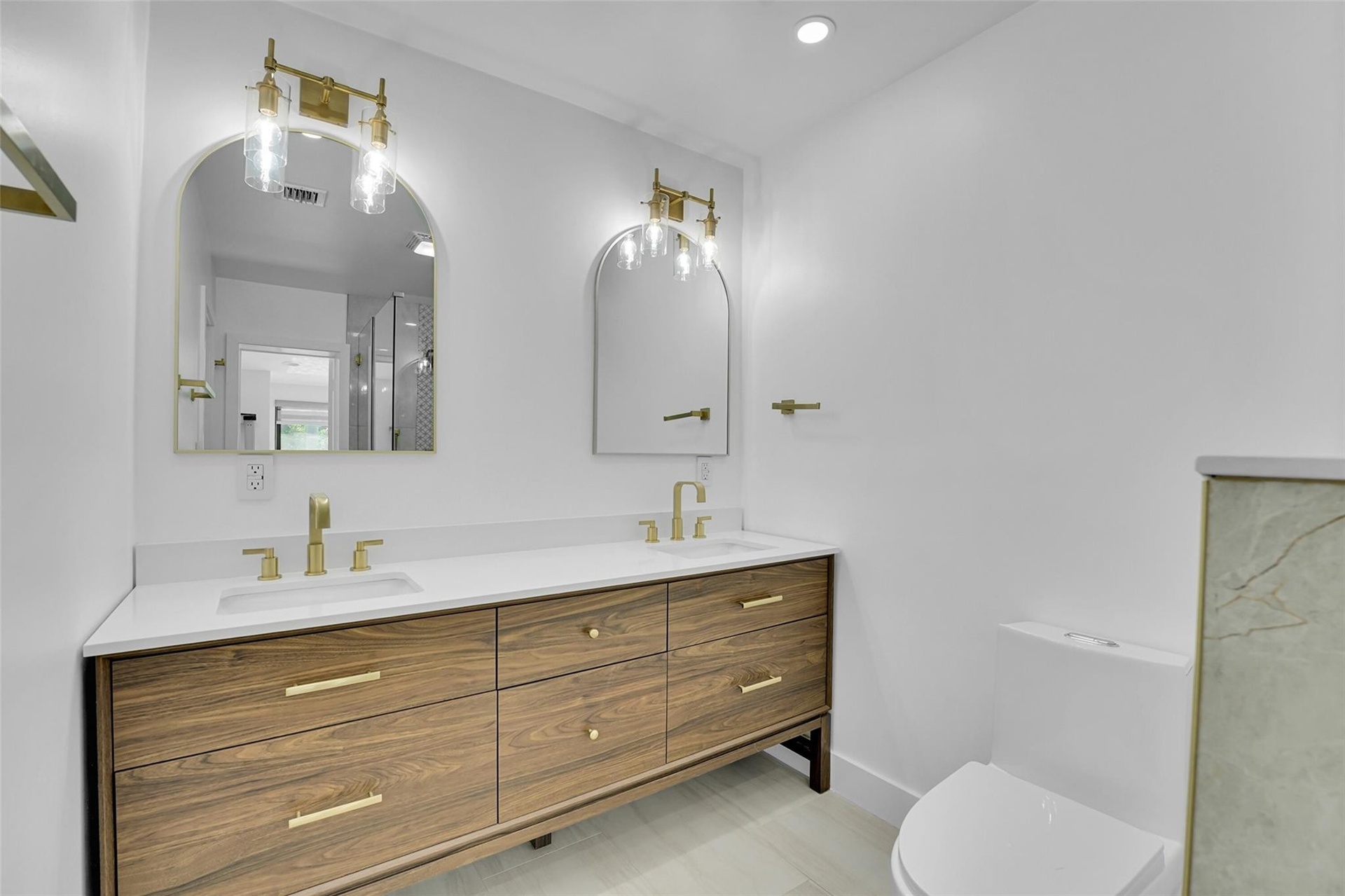 Dual vessel sinks, wood-finish vanity with drawer storage, polished brass fixtures, arched mirrors, and recessed lighting