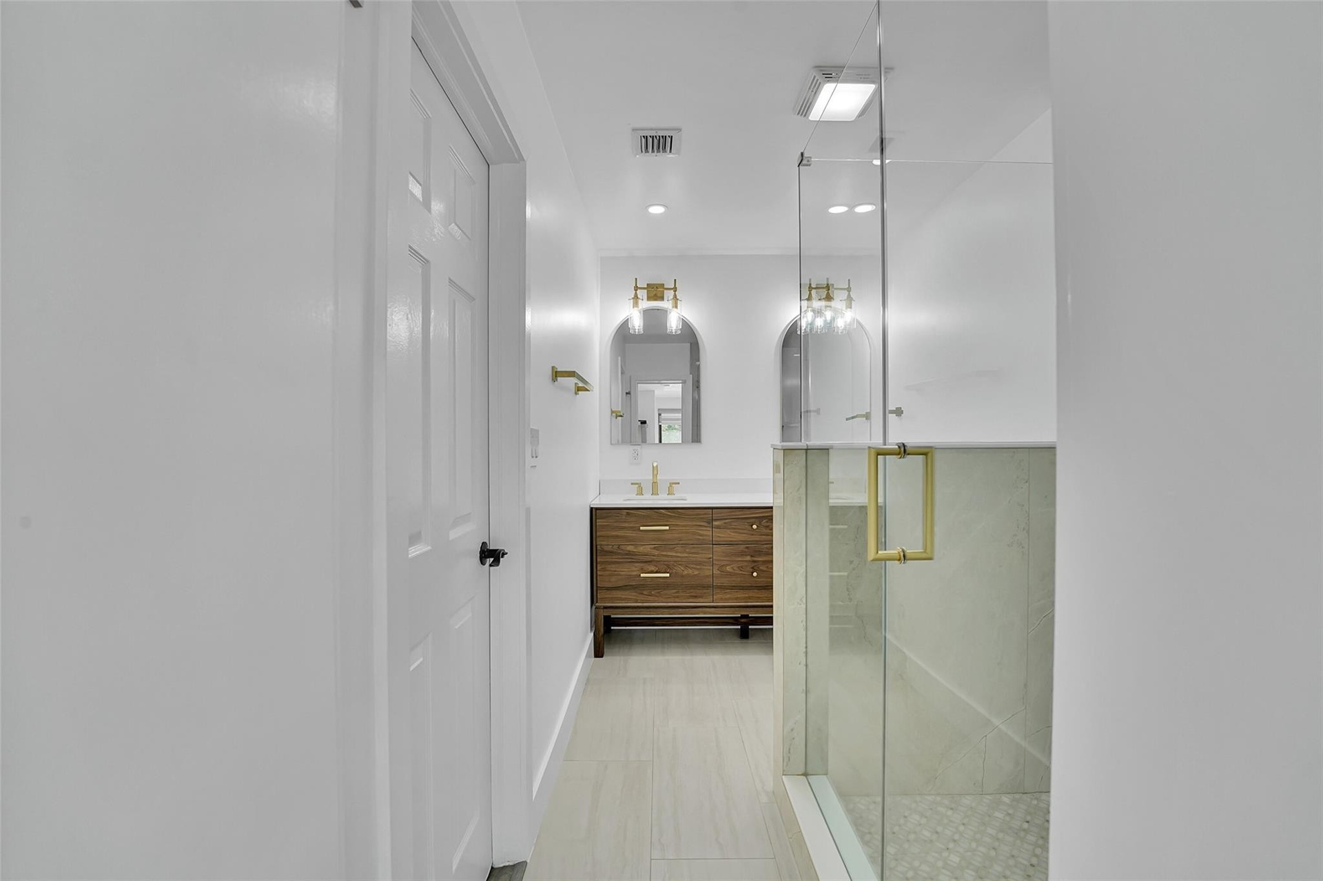 Glass-enclosed shower with gold-tone hardware, wood-finish vanity with white countertop, arched mirrors, and wall-mounted light fixtures