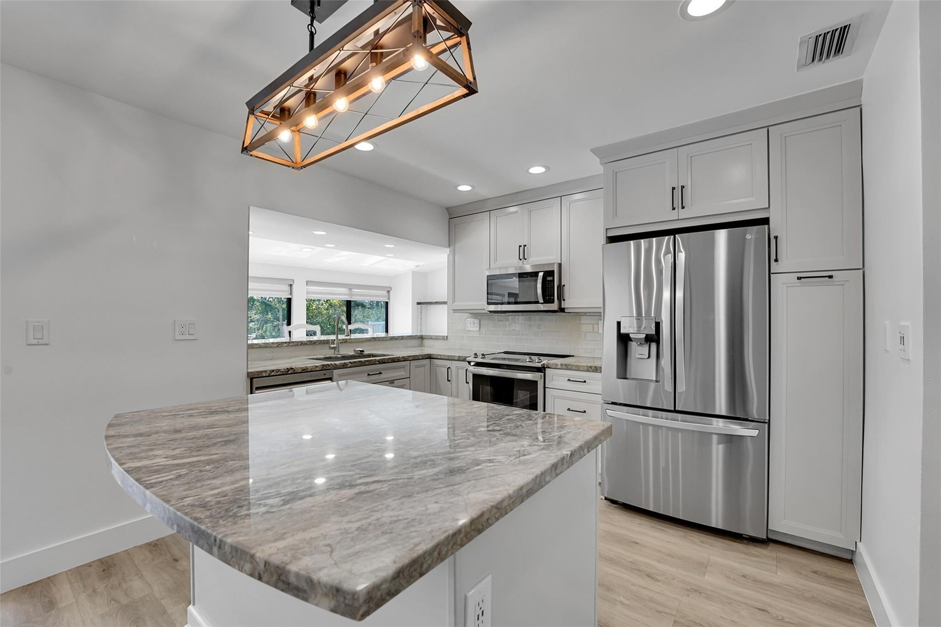 Kitchen featuring a stone-finish island with seating, a five-light linear chandelier, stainless steel appliances, light-toned cabinetry, and wood-fini