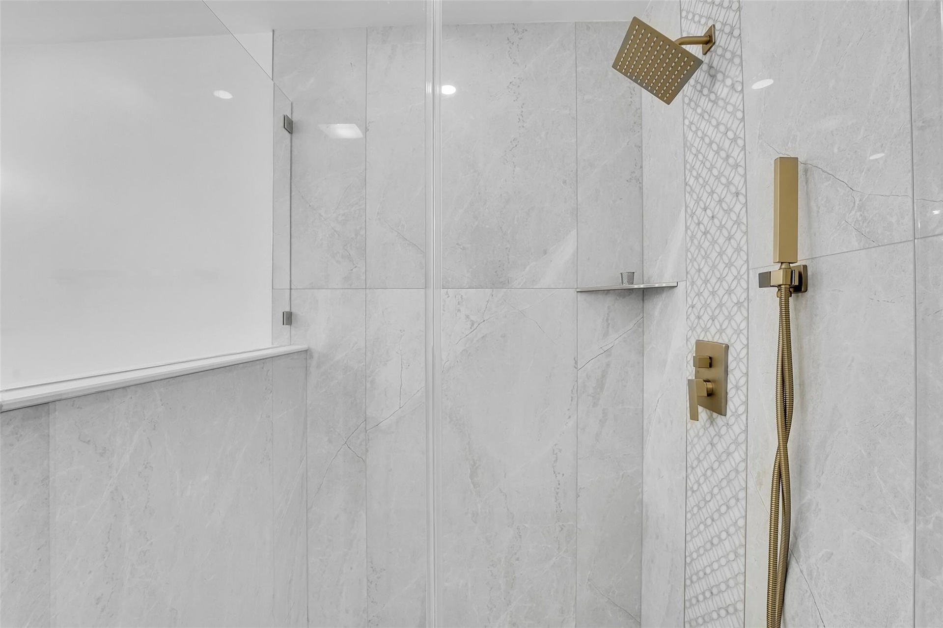 Shower enclosure with floor-to-ceiling glass panel, large format tile, decorative accent tile, built-in shelf, and a gold-toned shower system