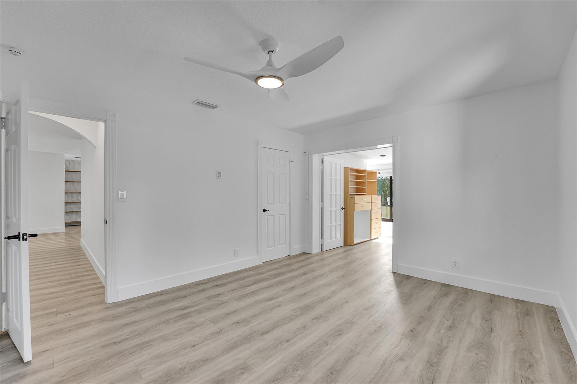 Light wood-finish flooring, white walls, modern ceiling fan with integrated lighting, and multiple doorways