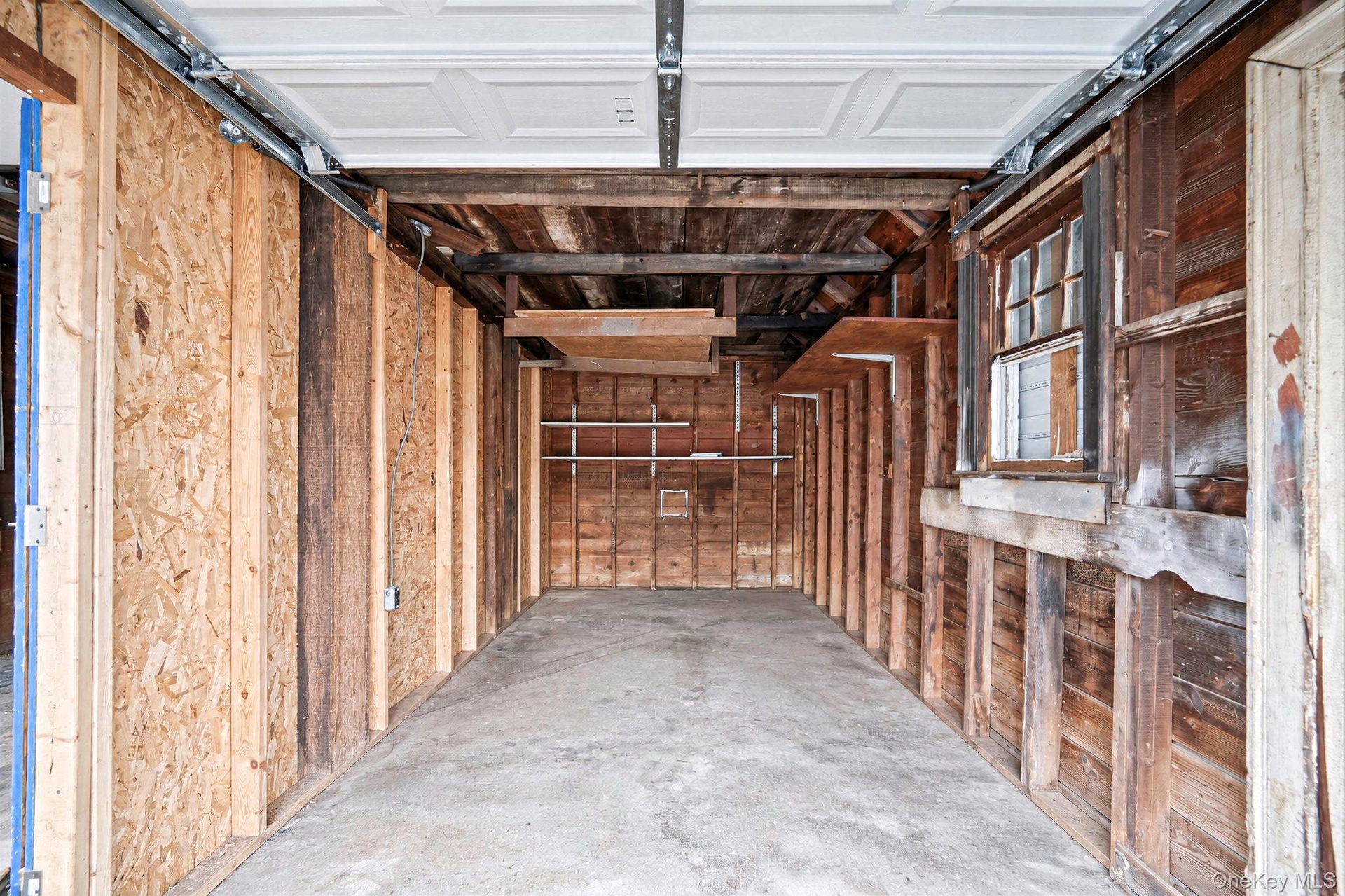 Enclosed garage featuring a concrete floor, exposed timber stud walls, integrated wooden shelving, and a multi-pane window