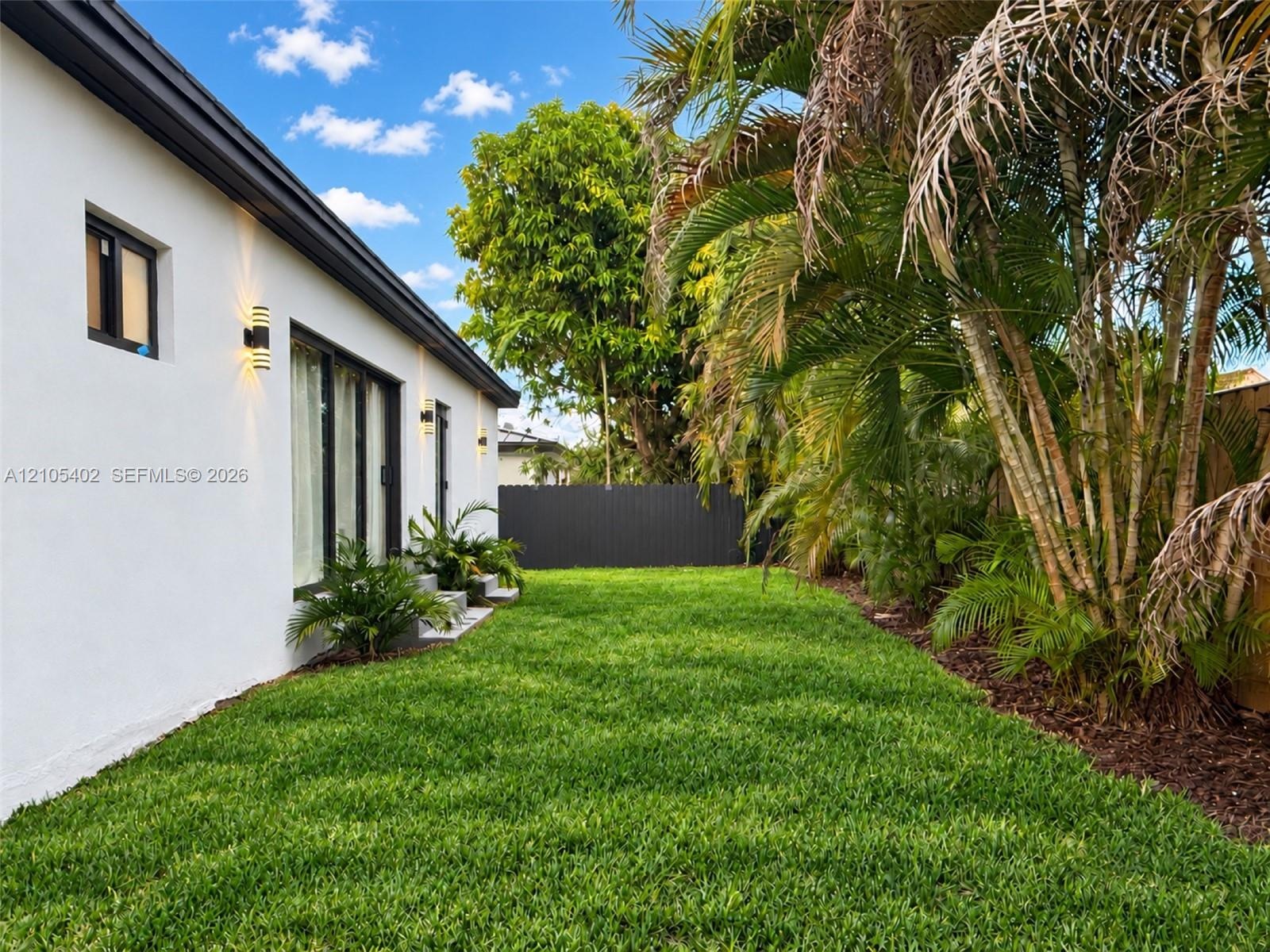 Exterior featuring white stucco siding, black-framed glass sliding doors, dark metal wall-mounted exterior lighting, manicured lawn, and mature palm t