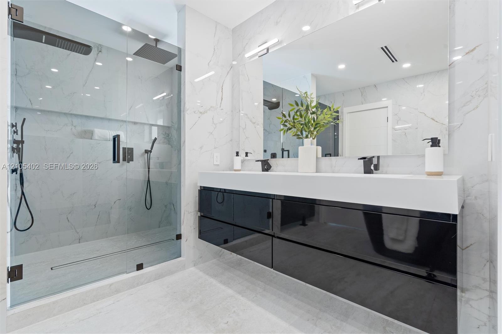 Floating high-gloss black vanity featuring a dual-sink white countertop, wall-to-wall mirror, glass-enclosed walk-in shower with stone-finish wall til