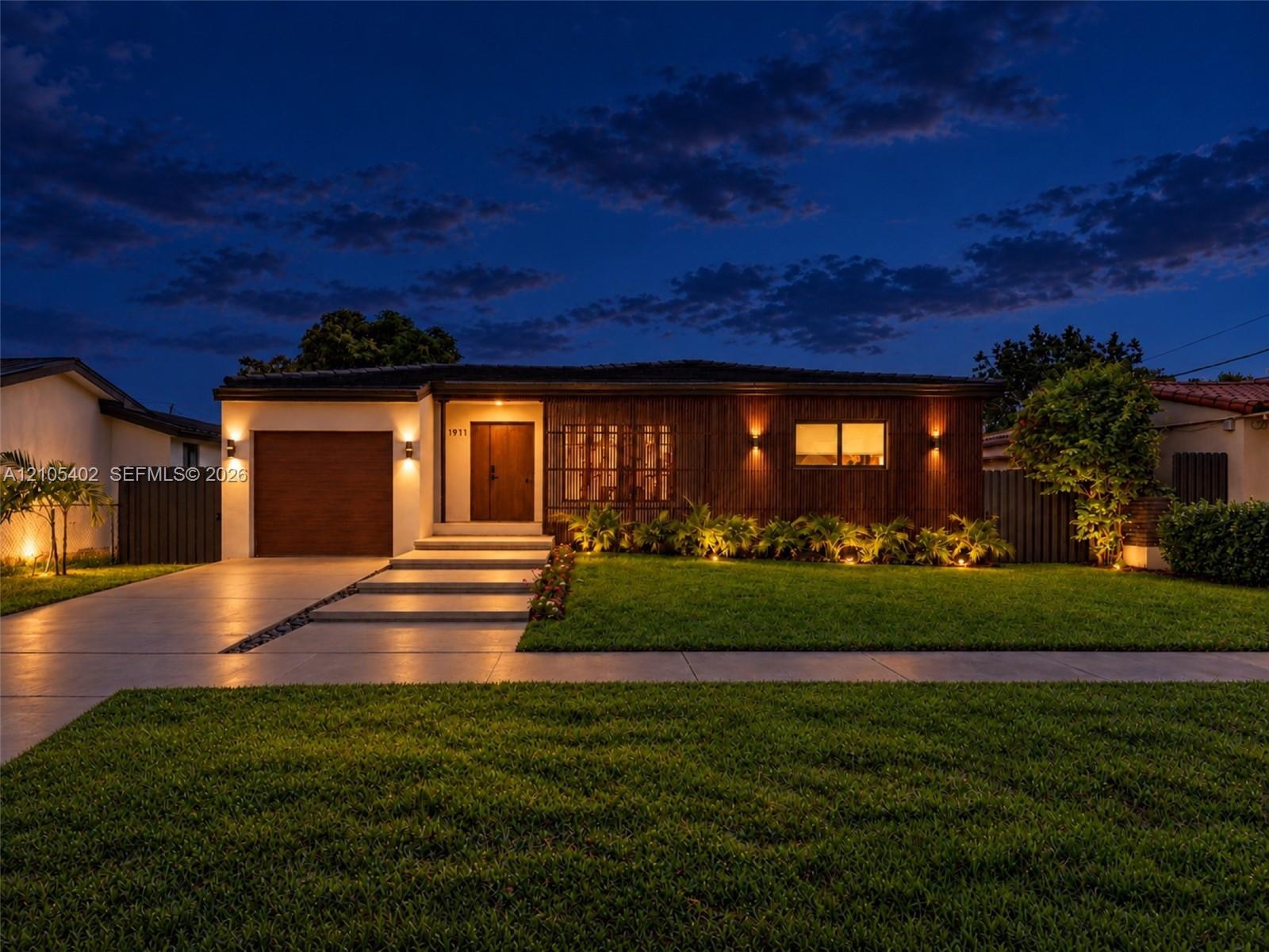 Modern single-story exterior featuring a wood-panel façade, integrated wall sconce lighting, solid wood front door, horizontal-slat garage door, and c