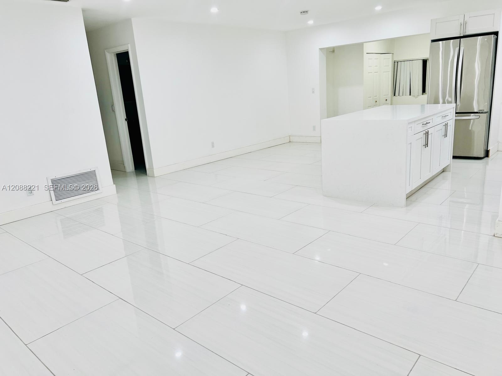 Large-format white tile flooring, central kitchen island with countertop, and stainless steel refrigerator