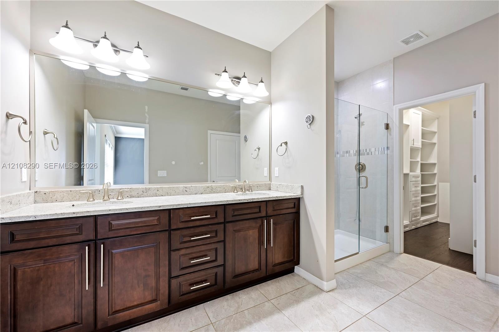 Double vanity featuring dark wood-finish cabinetry, stone-surface countertop, dual undermount sinks, and a large-format vanity mirror with dual light 