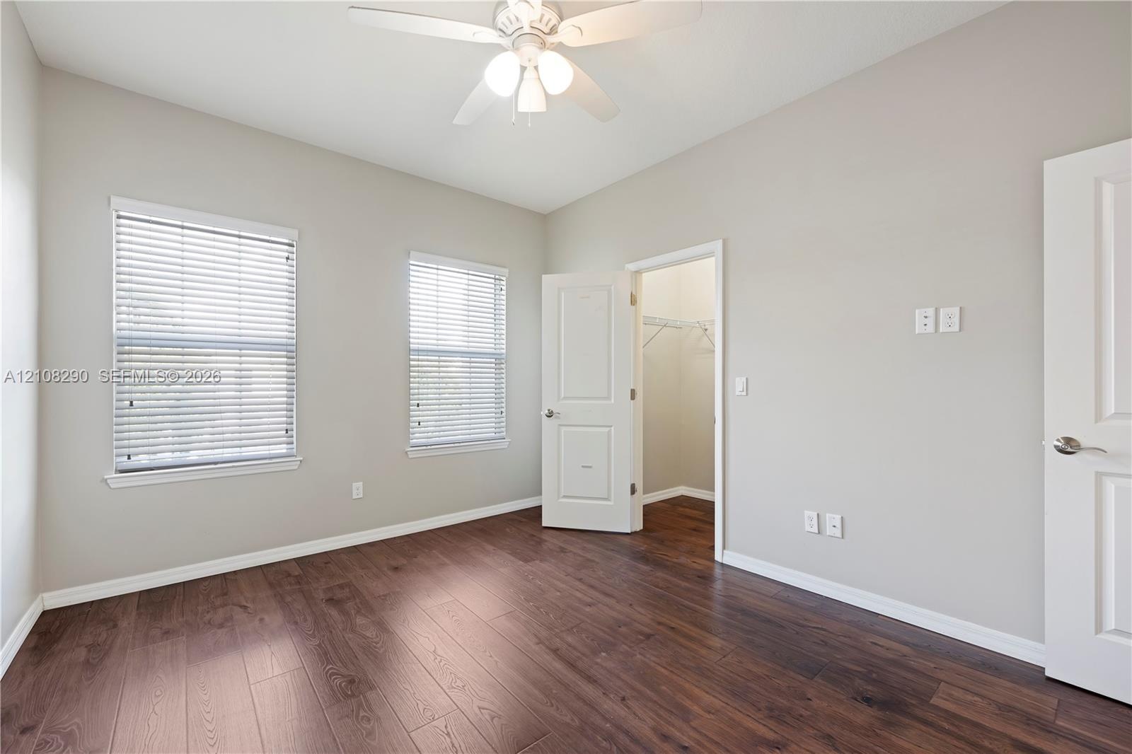 Interior space featuring wood-finish flooring, two windows with horizontal blinds, a ceiling fan with light fixture, and an open doorway to a walk-in 