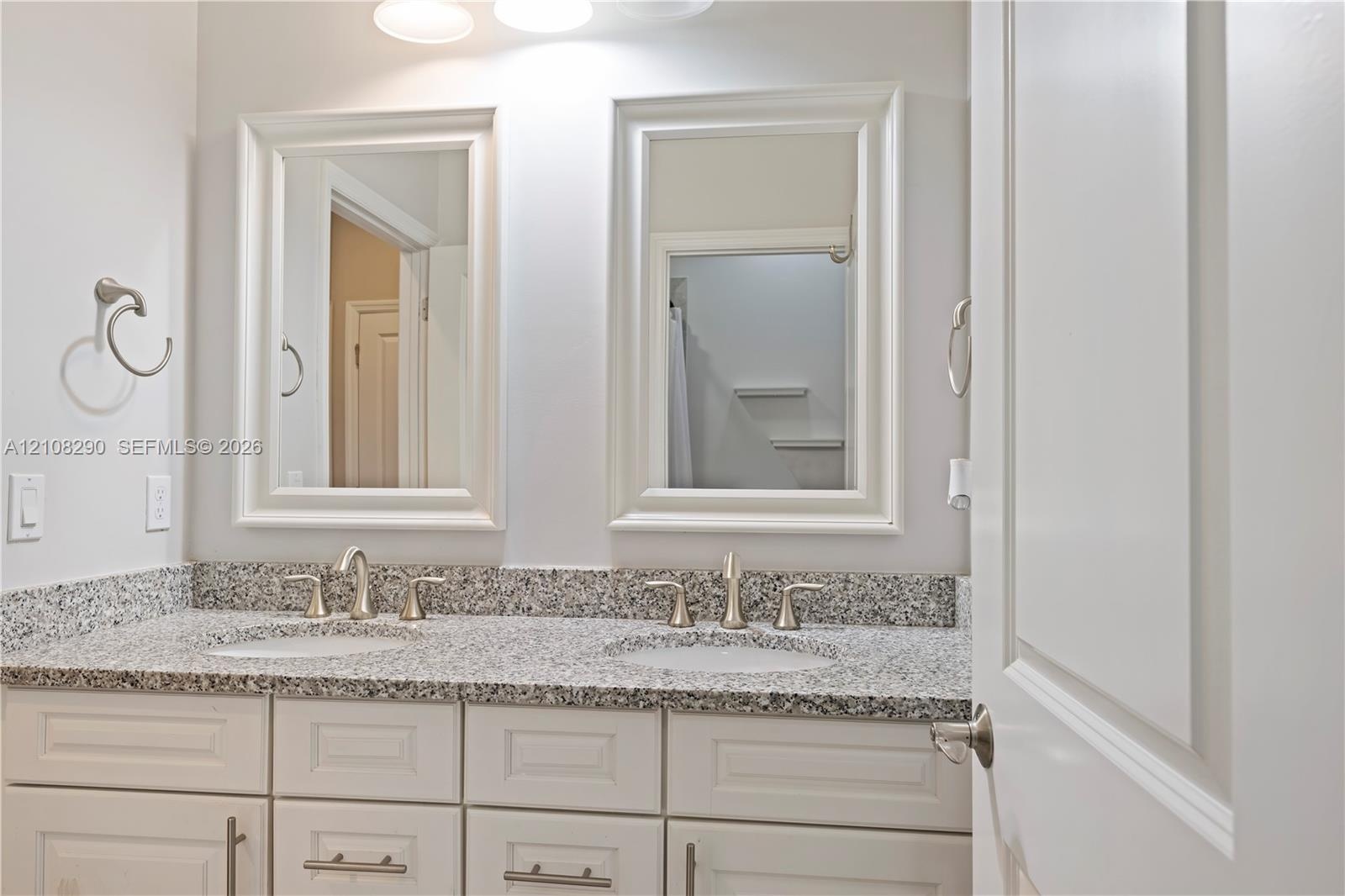 Dual-vanity cabinet featuring white shaker-style cabinetry, speckled stone countertop, integrated undermount sinks, and two framed wall mirrors with t