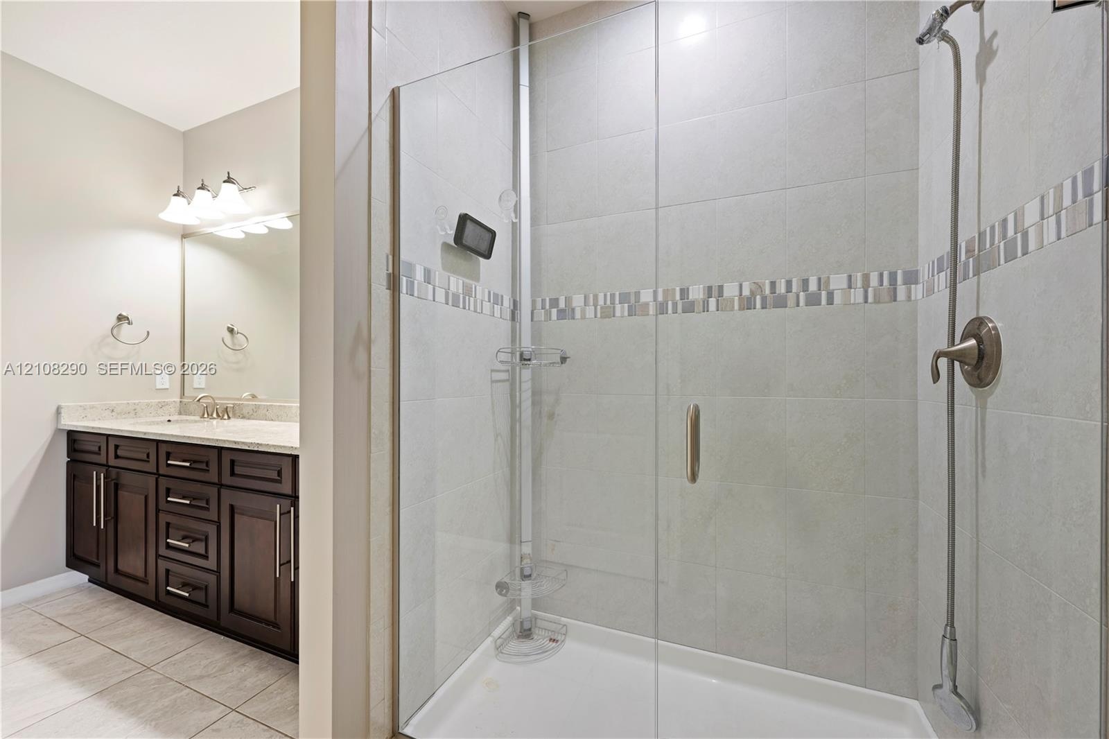 Walk-in shower with a glass enclosure, tile surround, and metallic fixtures, featuring a vanity with stone-finish countertop, dark wood cabinetry, and