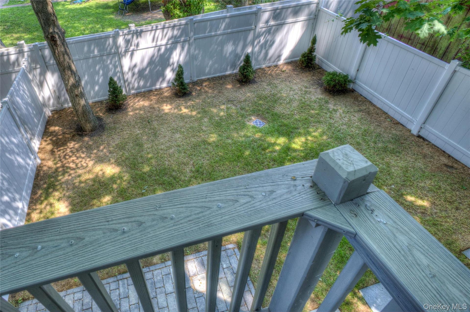 Fenced backyard featuring a wood-tone balcony railing, stone-finish paver patio, lawn area, and mature tree