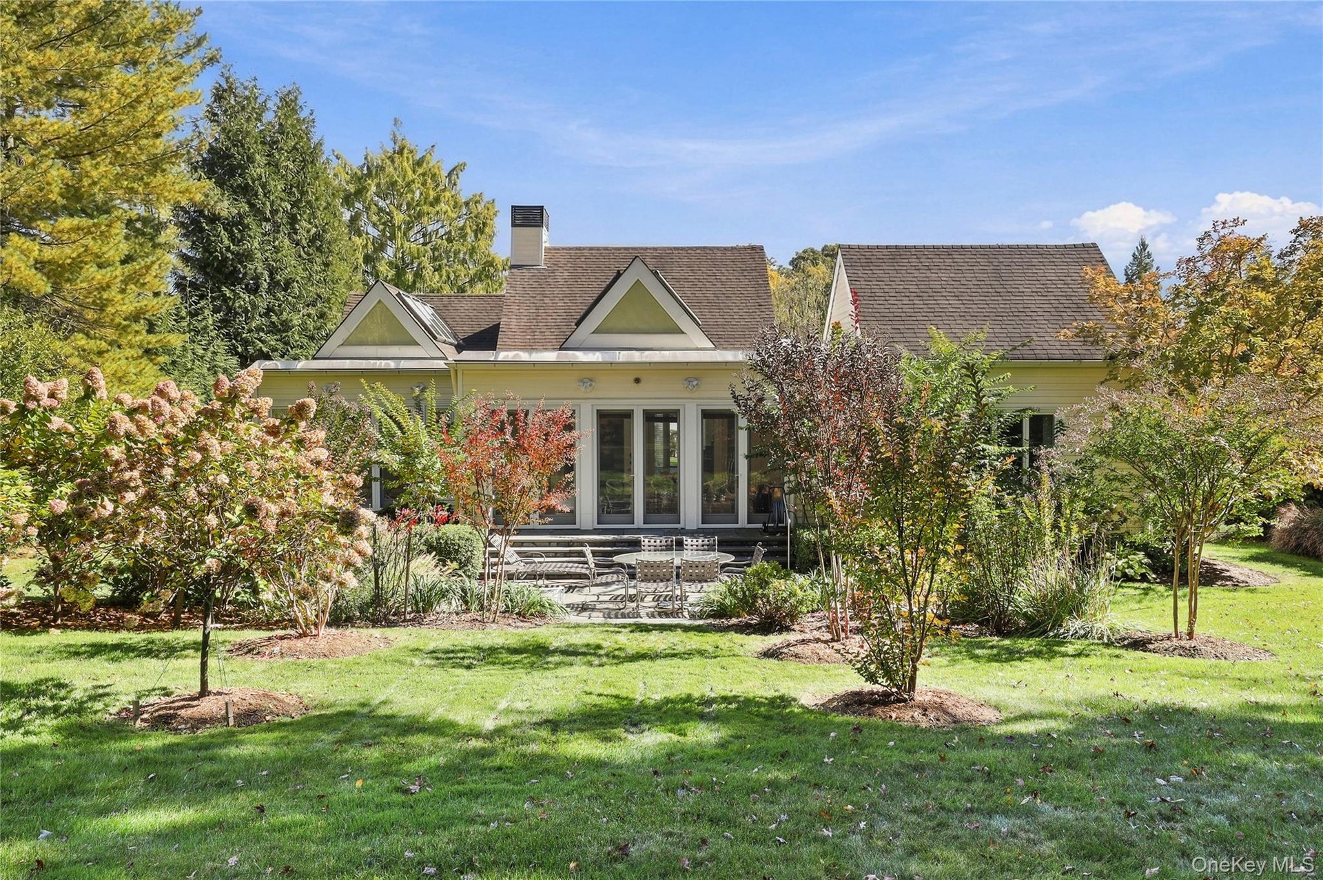 Rear exterior featuring a multi-gabled roofline, stone patio, glass-paned double doors, and established landscaping